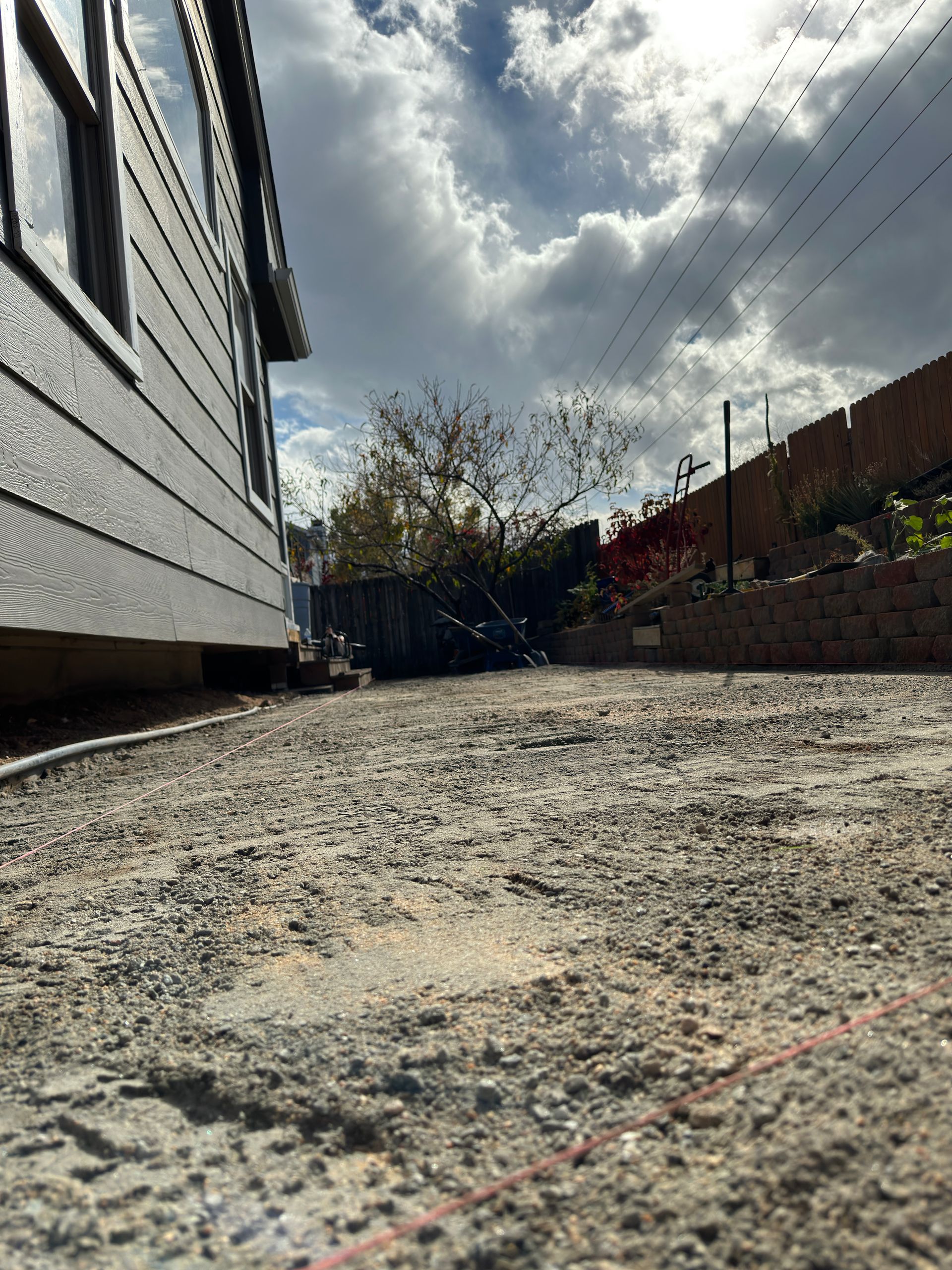 Gravel yard next to a grey house, with trees and a cloudy sky in the background.