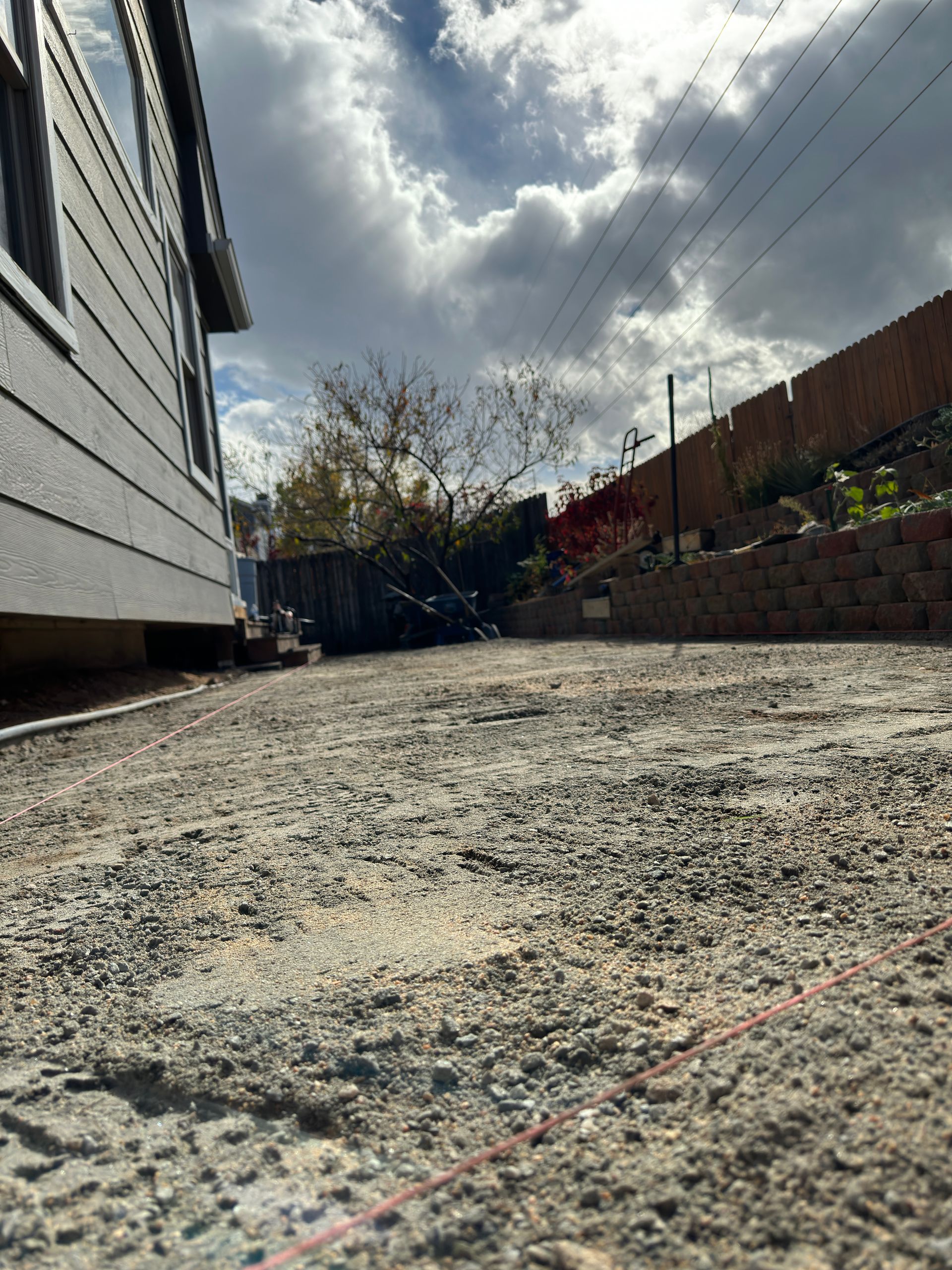 Gravel yard next to a gray house, with a fence and a retaining wall in the background under a cloudy sky.