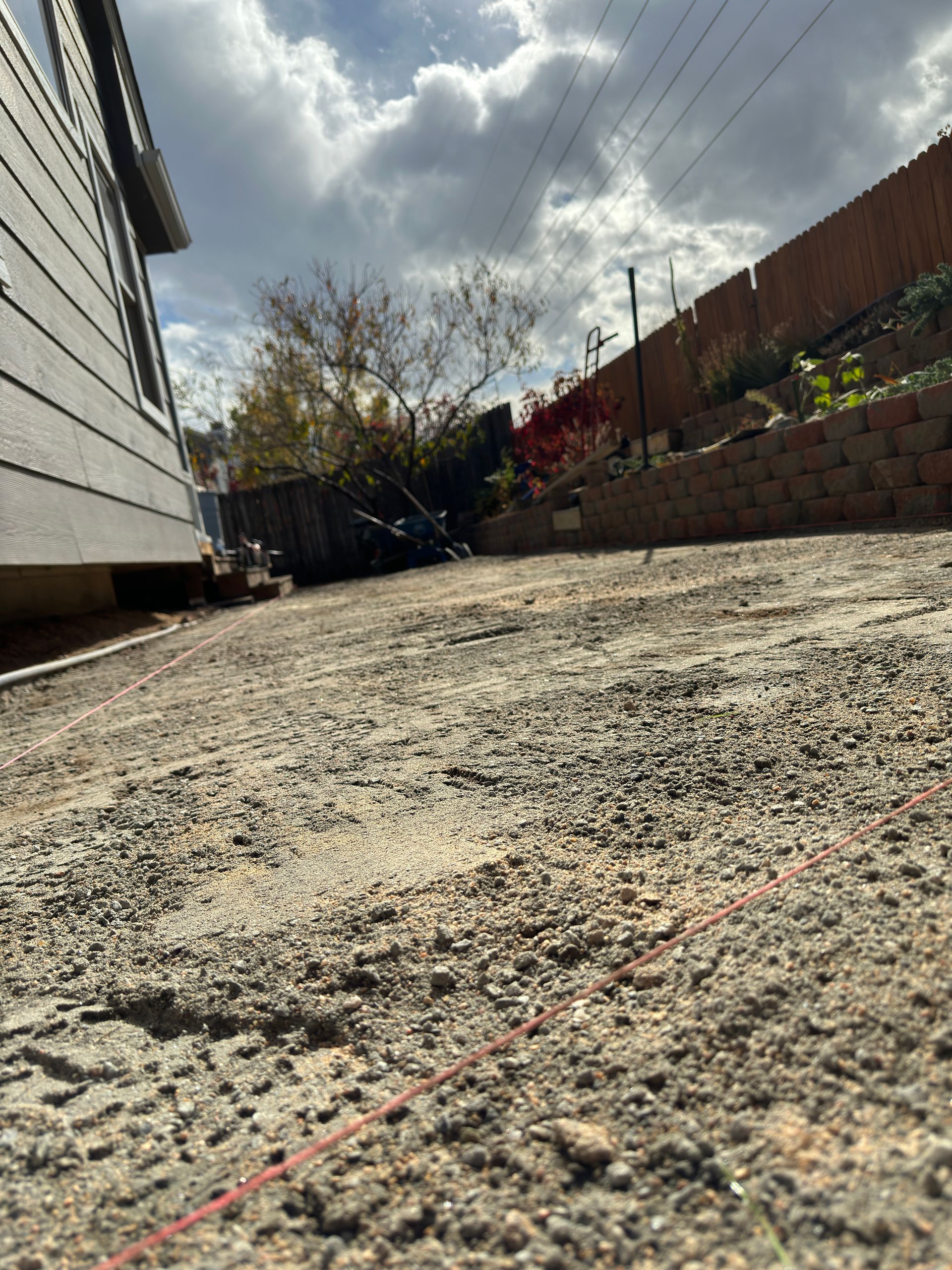 Gravel driveway with a house on the left, a wooden fence on the right, and a cloudy sky.