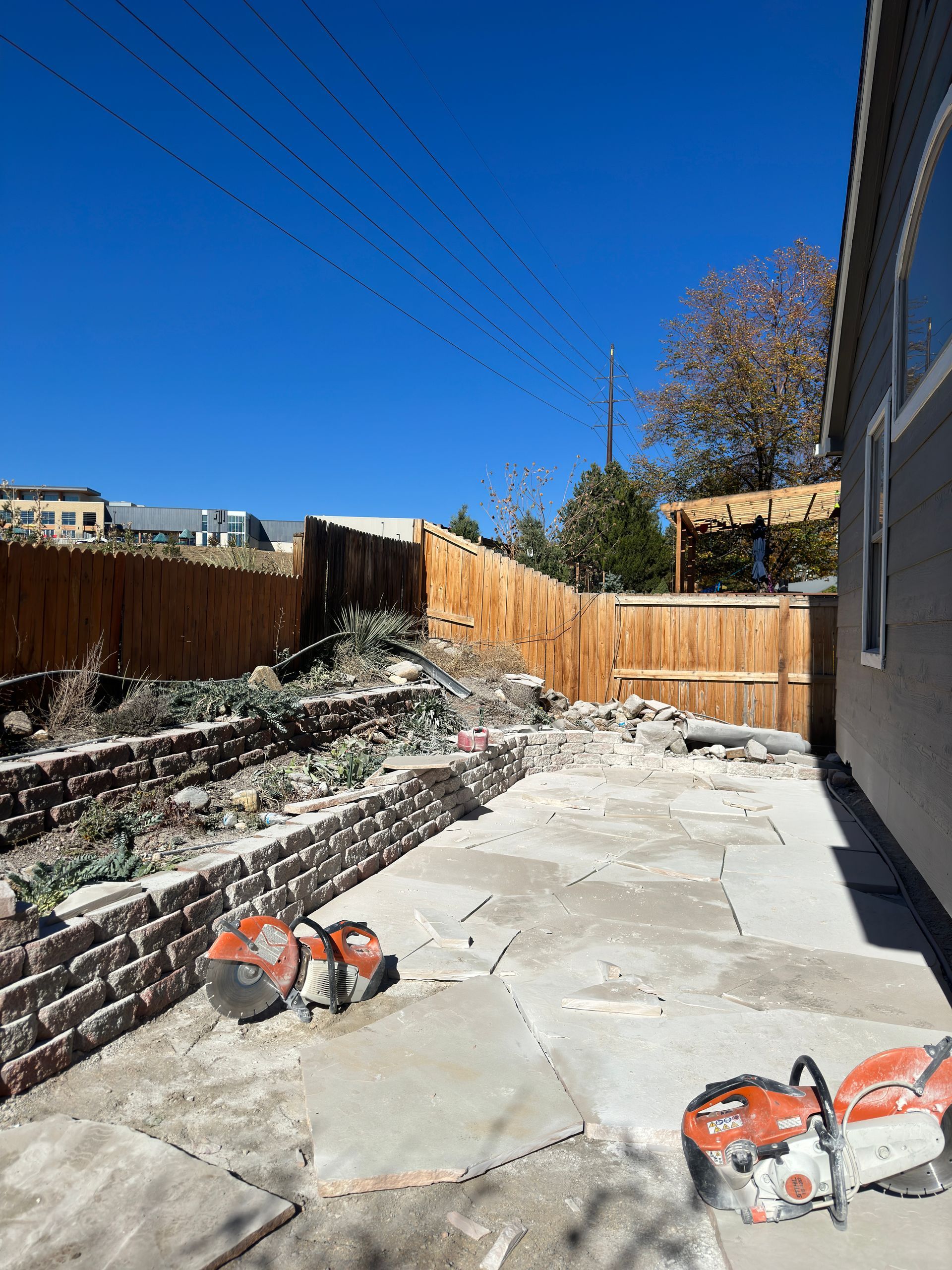 Backyard construction scene; two red saws on patio, brick retaining wall, wooden fence, clear blue sky.