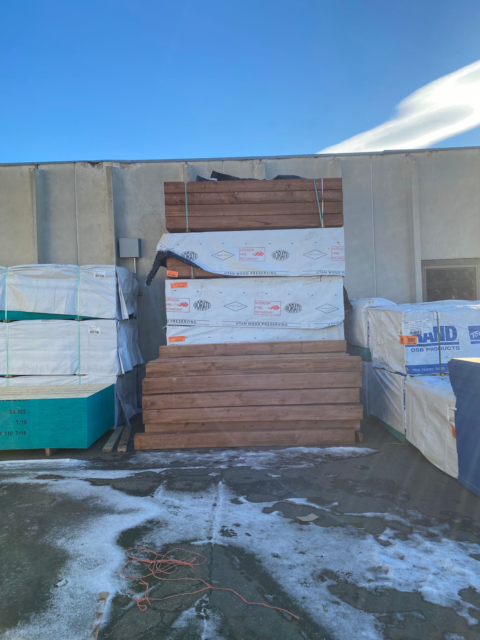 Lumber stacked outdoors in a snowy area against a gray building, with a clear blue sky above.
