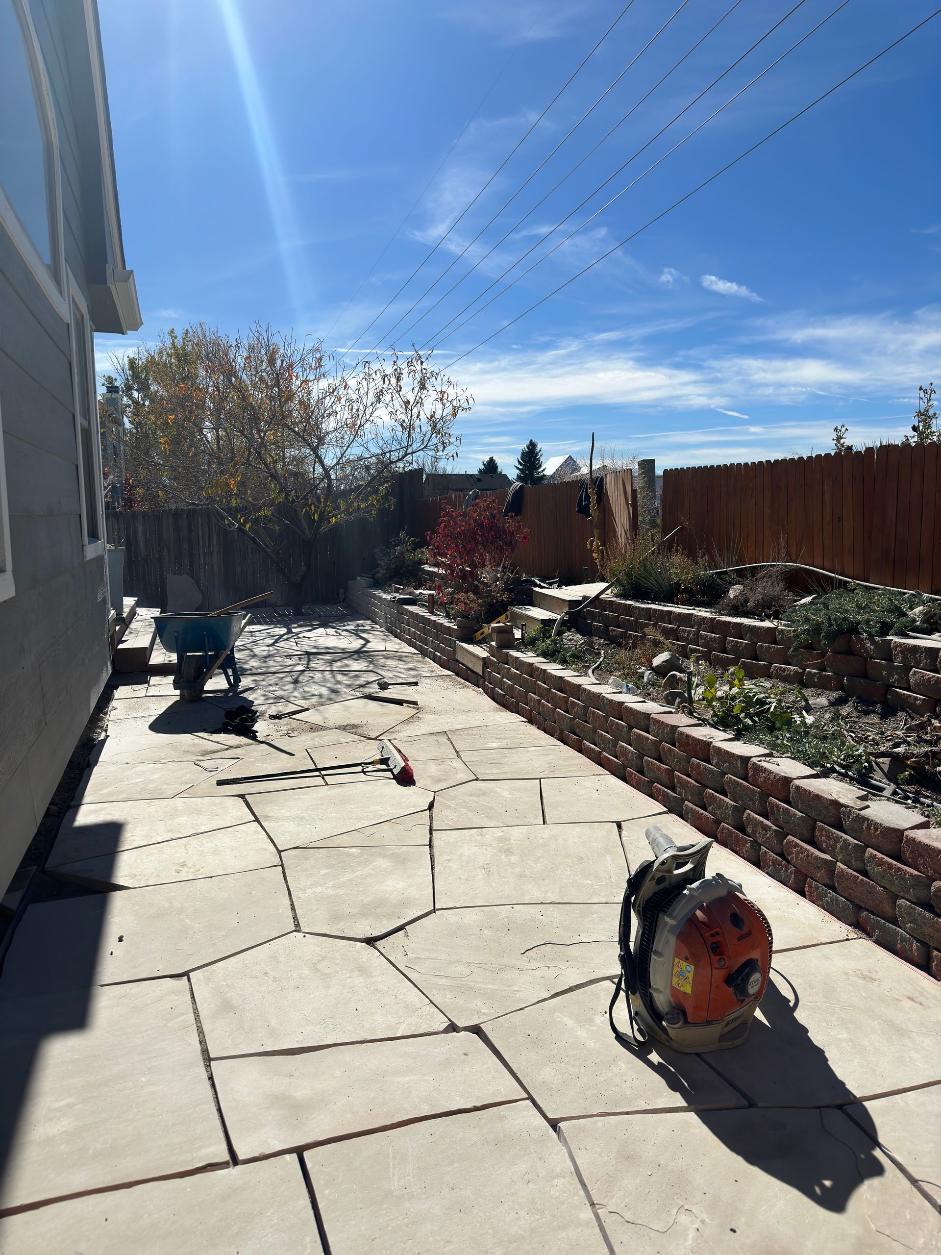 Backyard patio with flagstone, retaining wall, and leaf blower under a blue sky.