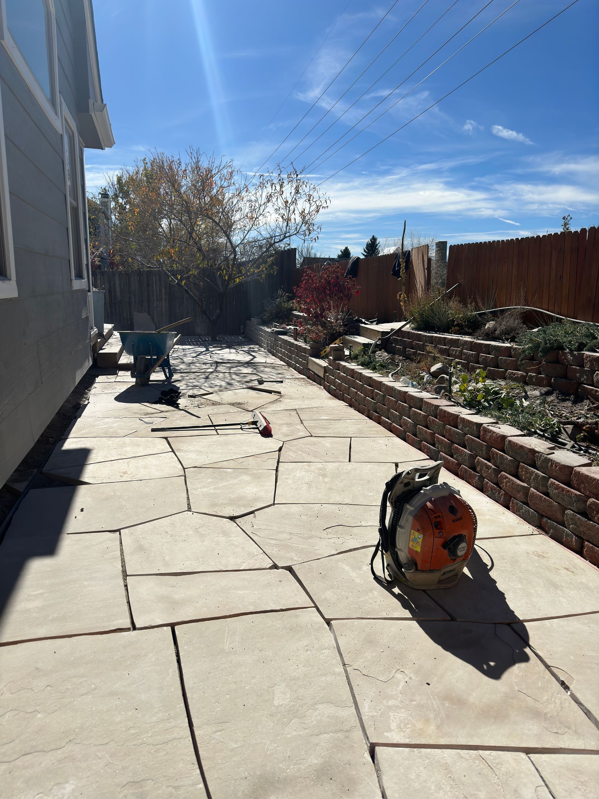 Patio with stone pavers, brick retaining wall, and leaf blower in a yard on a sunny day.