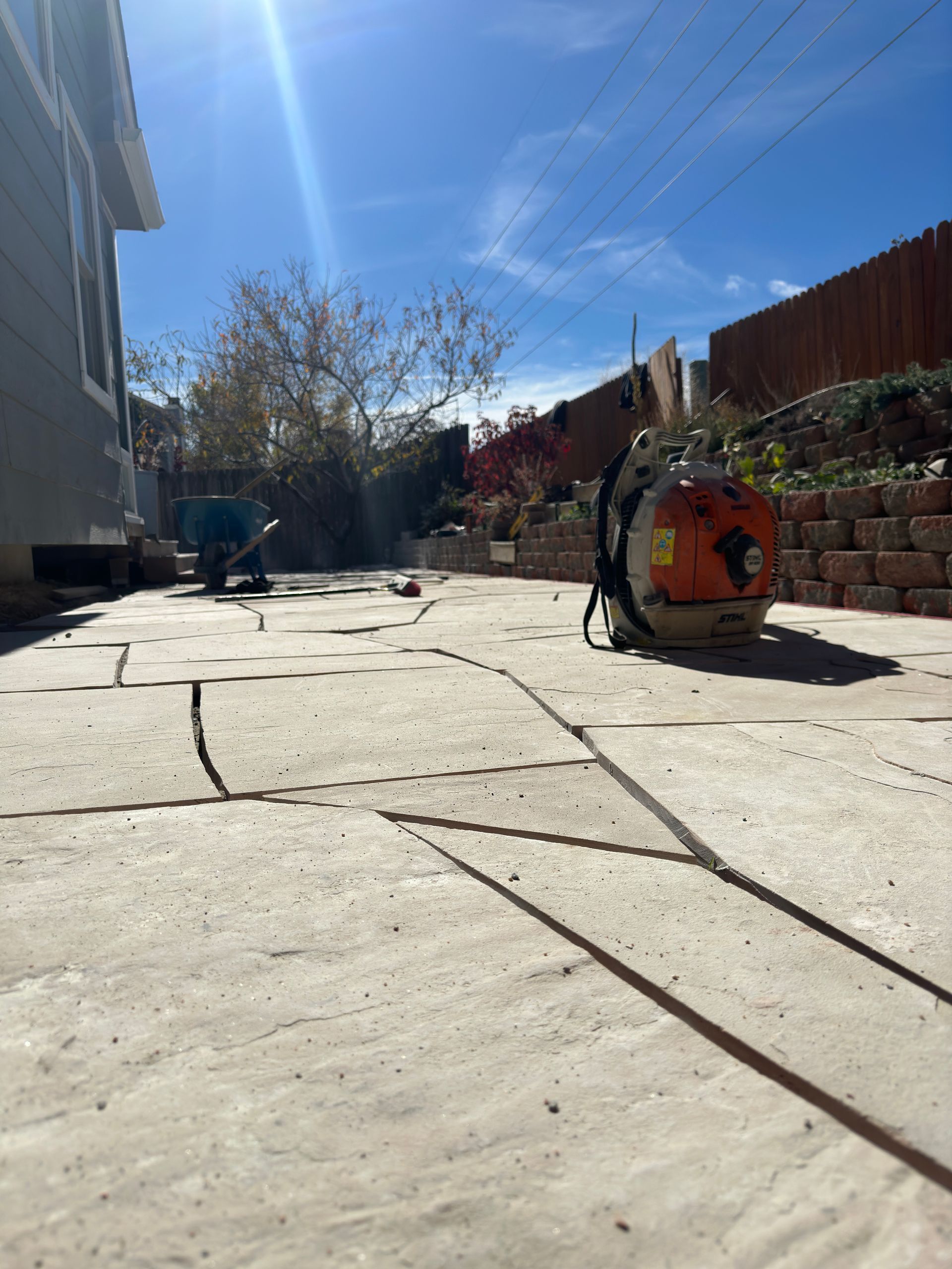 A sunny backyard patio with a leaf blower. A house and red fence are in the background.