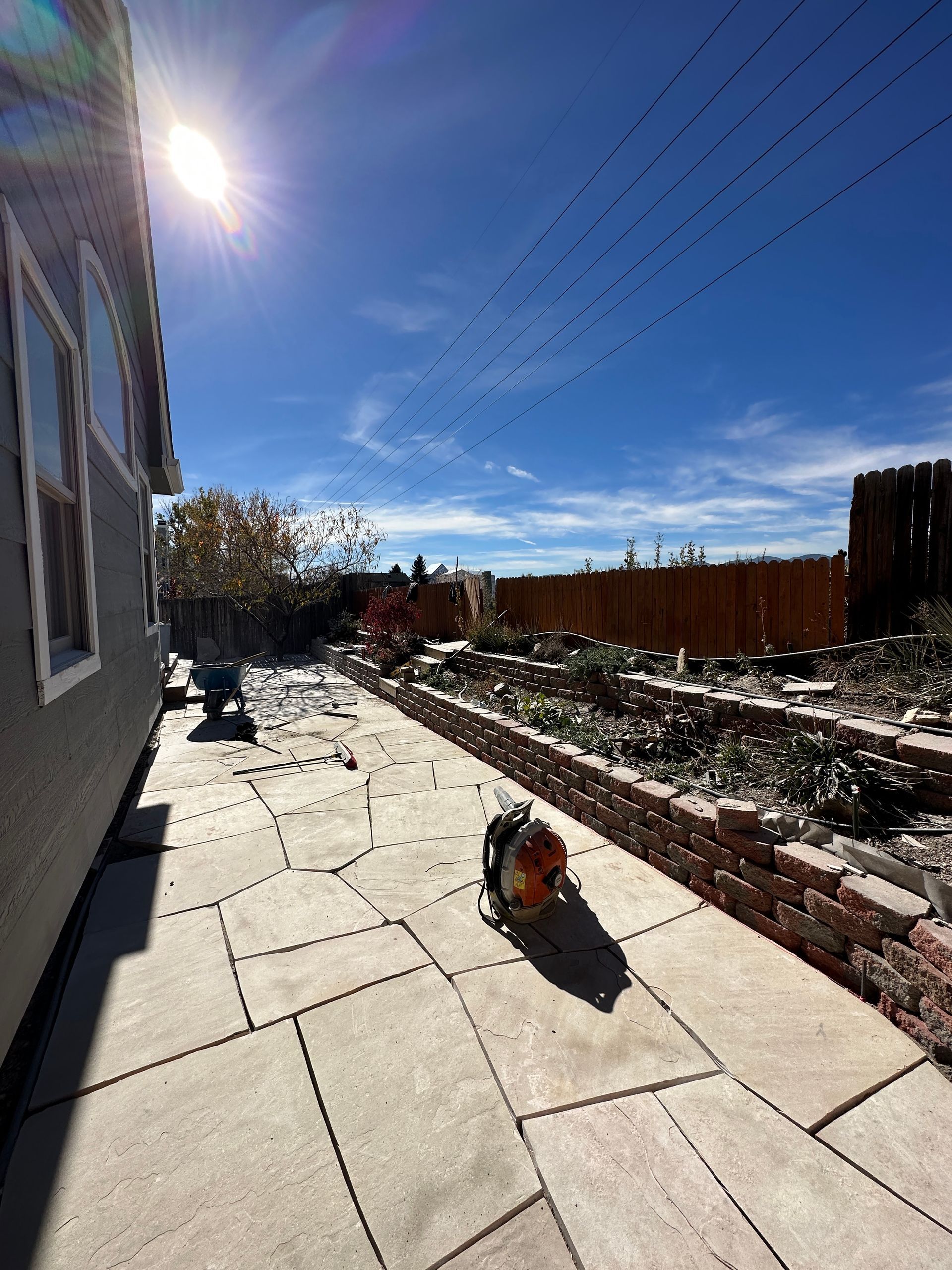 Backyard with stone patio, brick-lined garden, and hedge. Bright sun, clear blue sky.