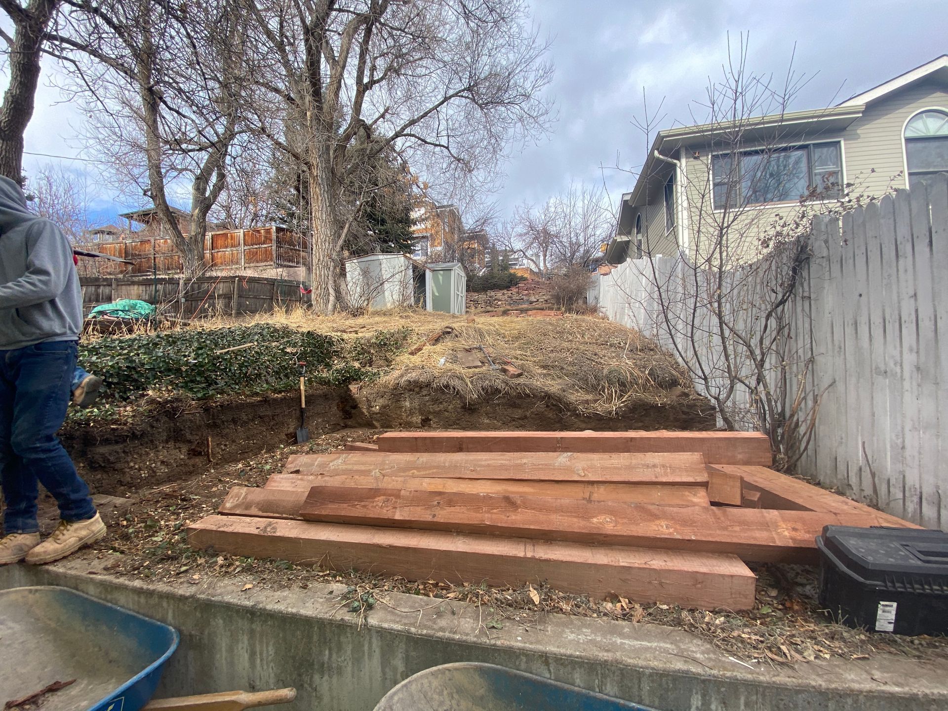 Person near stacked red timbers, possibly a retaining wall. Dirt hill in the backyard with some dead foliage, overcast sky.