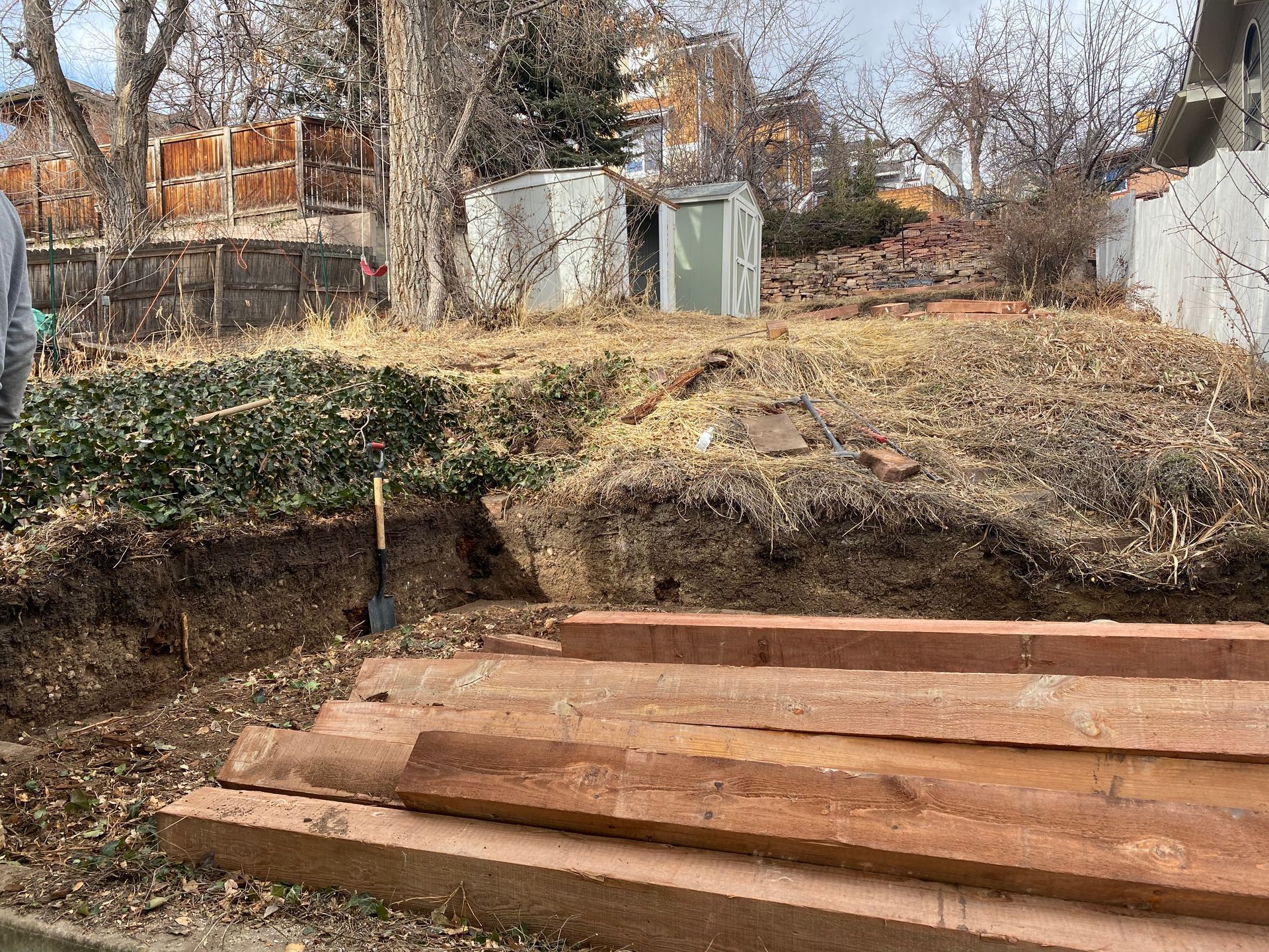 A sloped backyard with a small retaining wall and wooden planks. Shovel, dead grass, and a shed are visible.