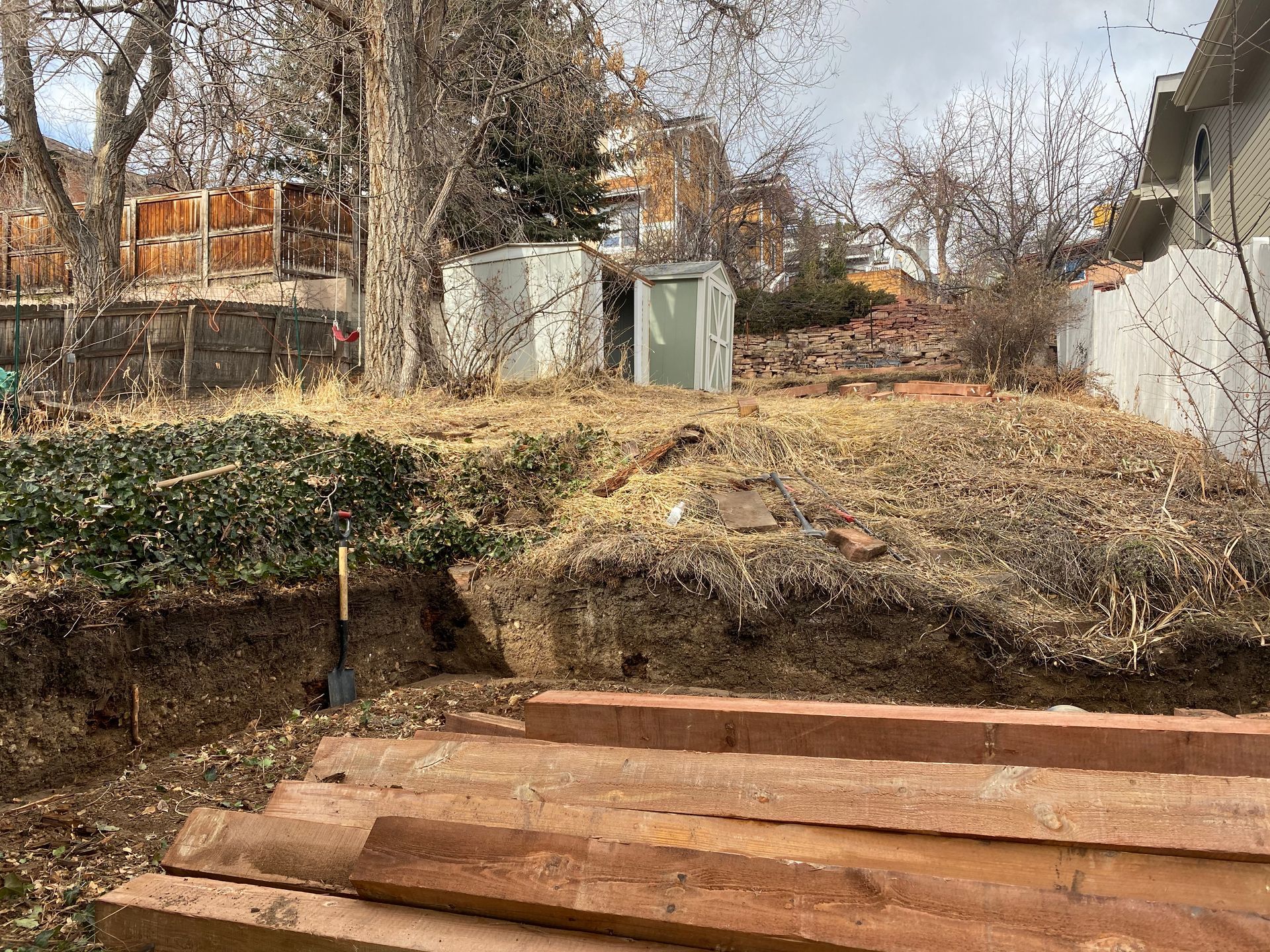 Brown wooden steps lead to a sloped, overgrown yard with two sheds and a shovel.