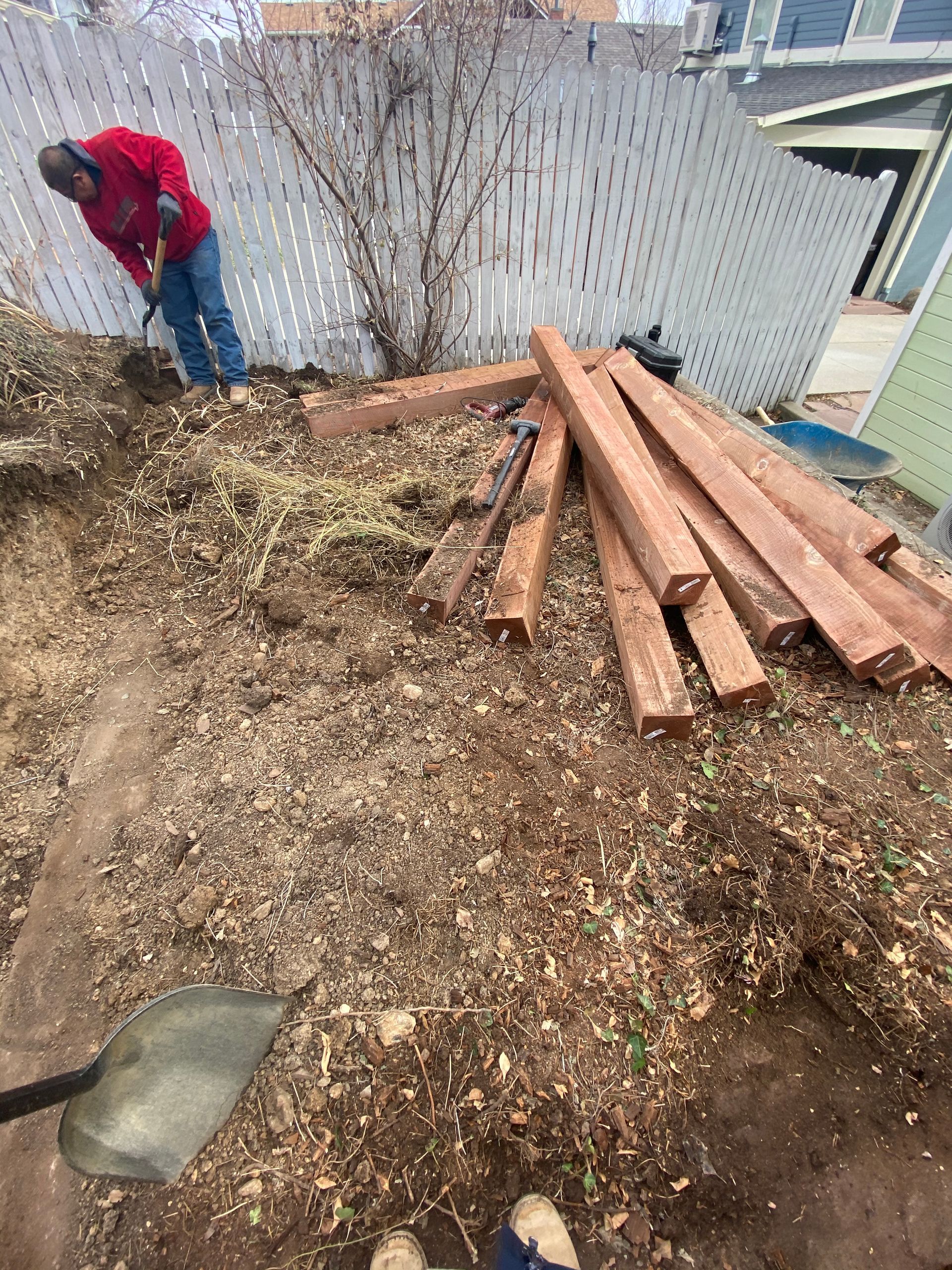 Man in red jacket shovels dirt, wood planks piled nearby, against a white fence.