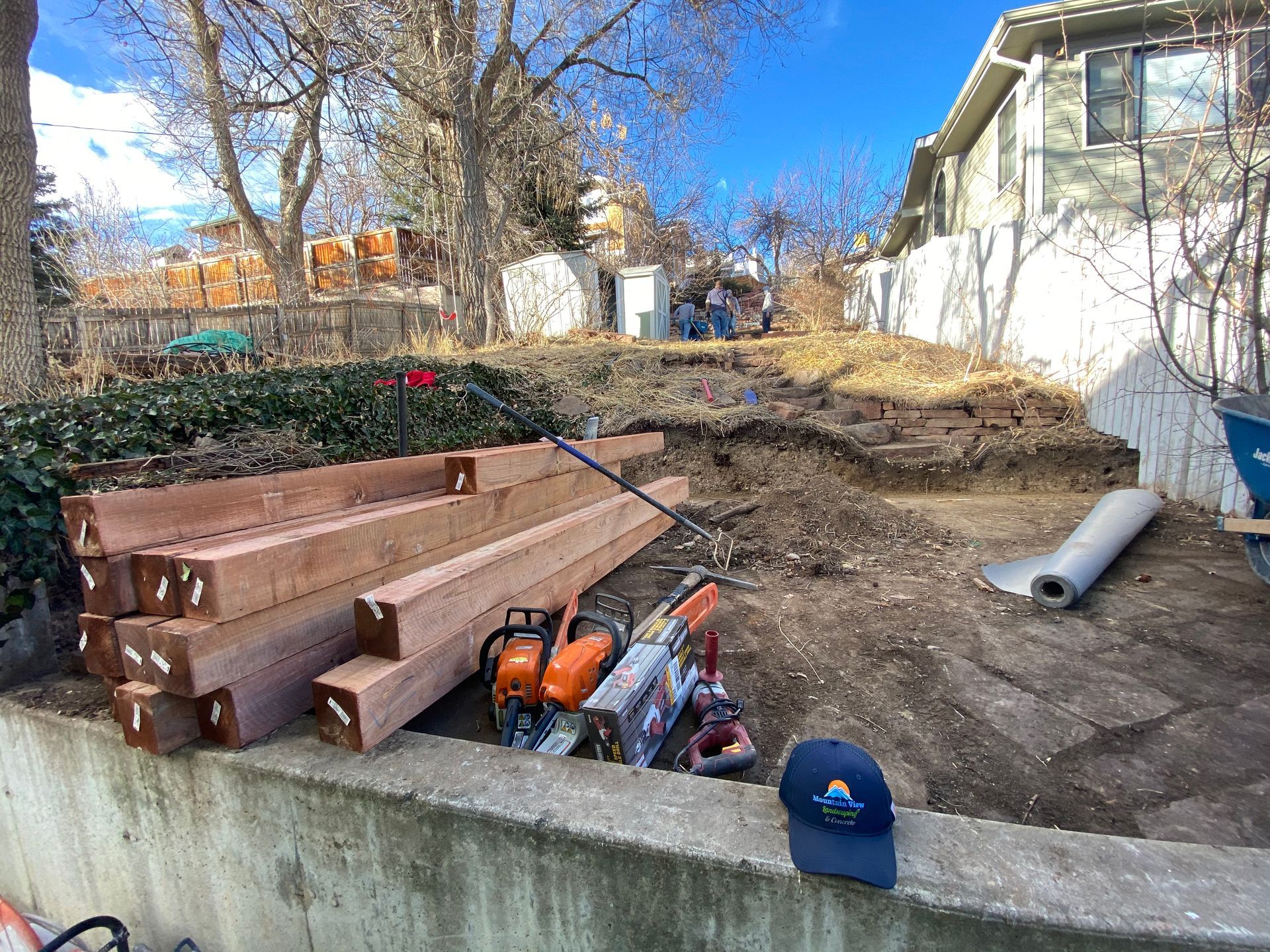 Construction site with stacked lumber, tools, and excavated hillside; a house and a blue sky are in the background.