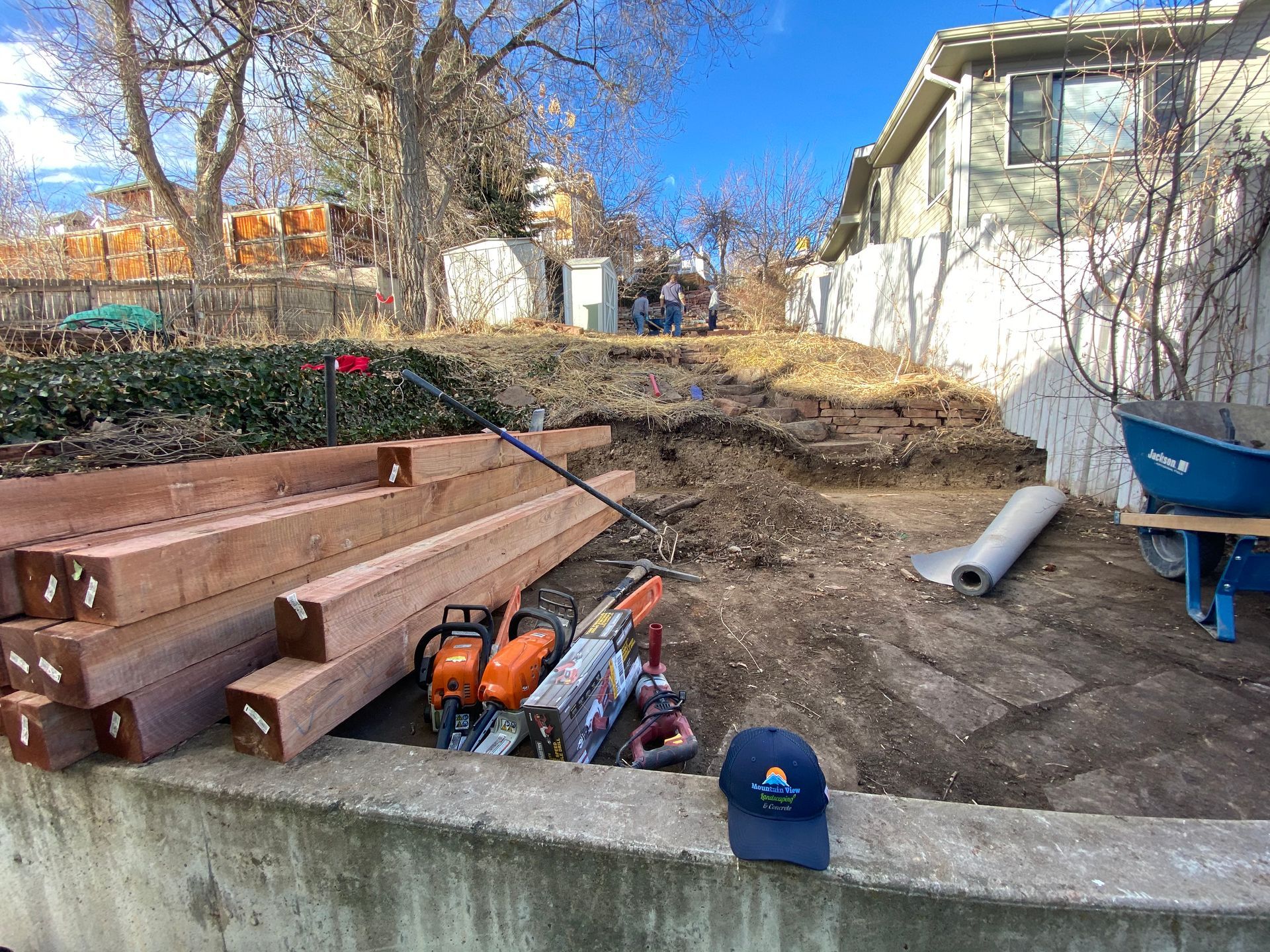 Construction site with stacked lumber, chainsaw, dirt hill, and a blue cap.