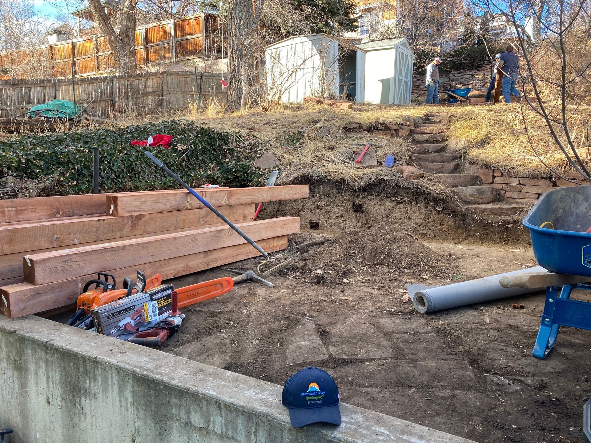 Backyard landscaping: lumber, chainsaw, dirt hill with steps, people working, blue wheelbarrow, and a blue cap.