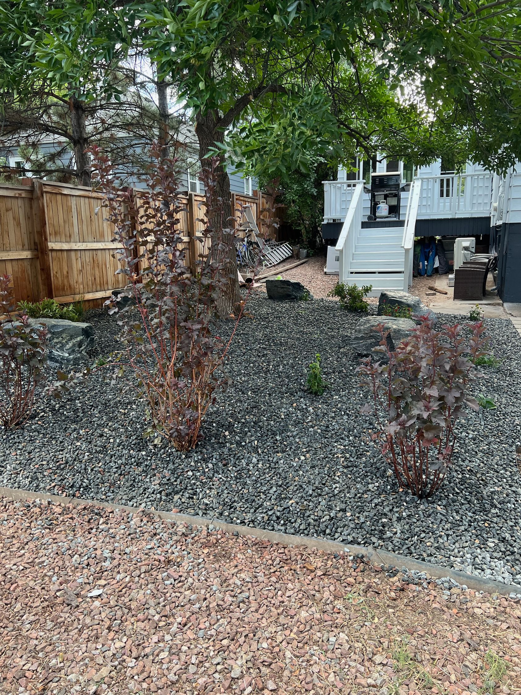 A landscaped garden bed with dark gravel, red-brown plants, and a wooden fence; a house with a deck is in the background.