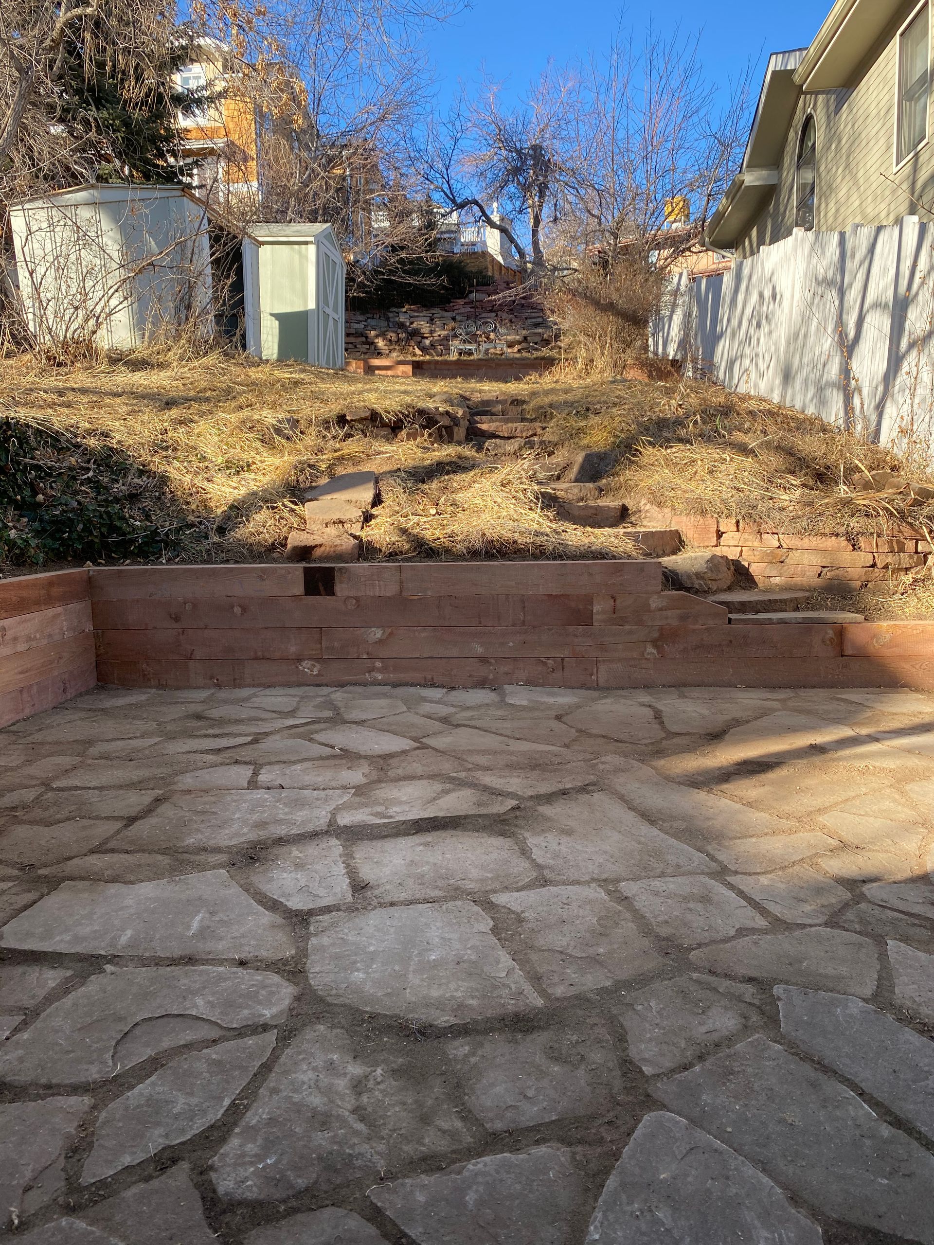 Stone patio in front of a hillside with wood retaining walls and steps leading up to a shed.