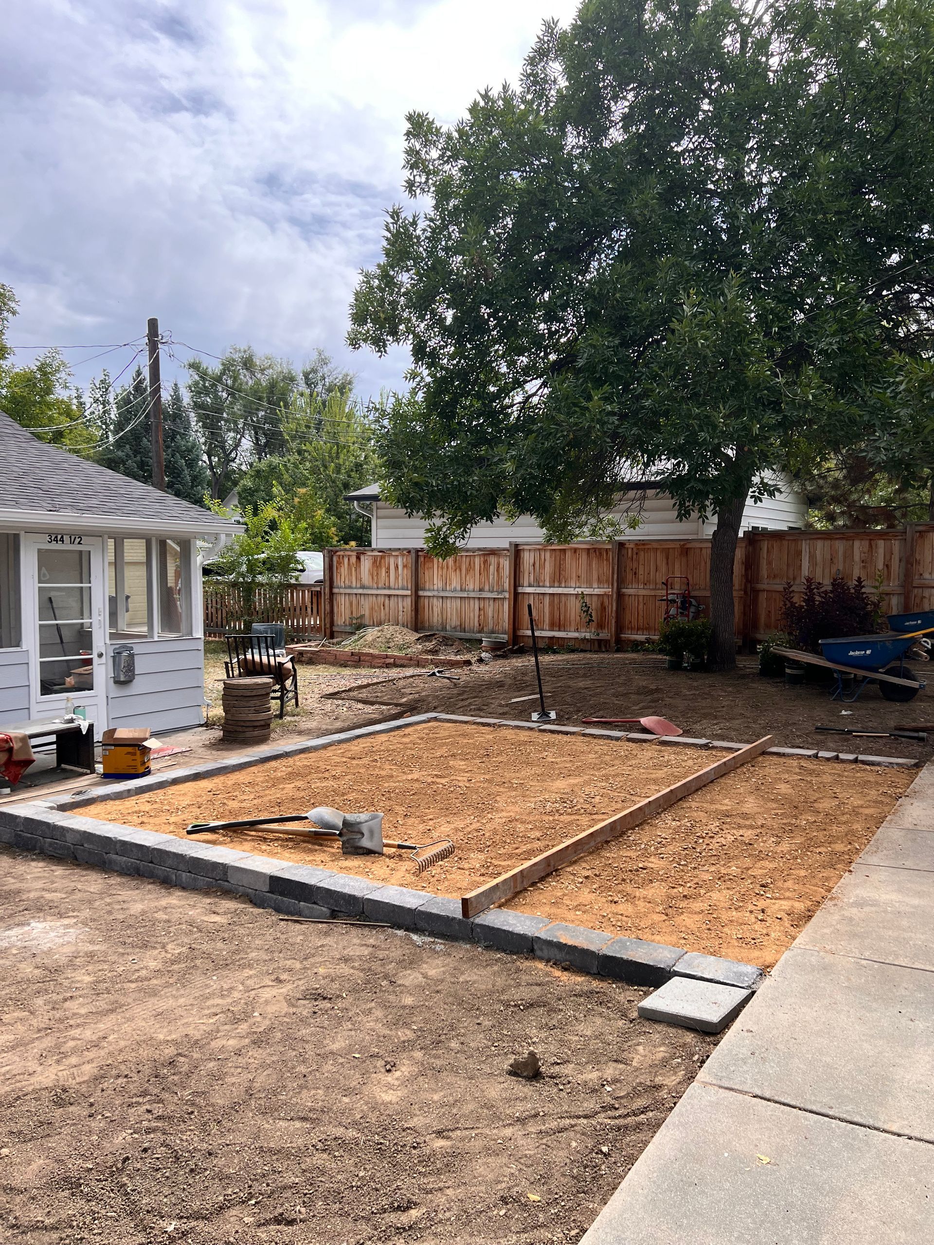 Backyard with wood-framed garden beds filled with mulch, bordered by paving stones. A fence and large tree are in the background.