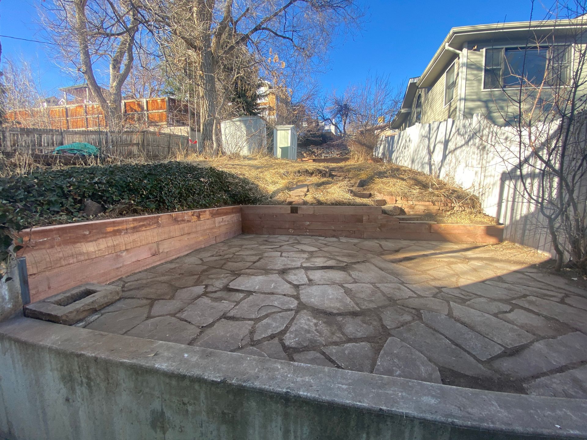 A backyard with a stone patio, retaining walls, and a hill leading to a fence and house under a blue sky.