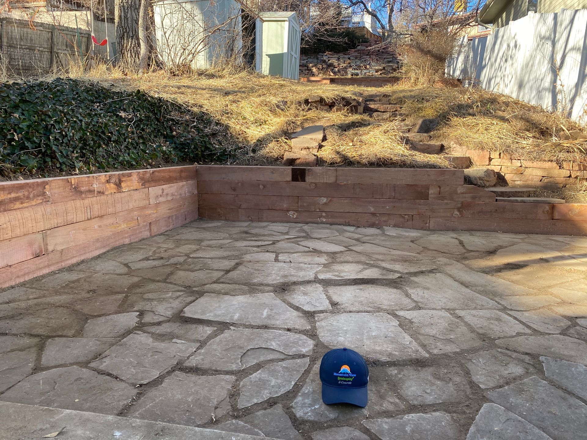 Stone patio enclosed by brick walls, leading to a sloped, overgrown hillside. A hat sits on the patio.