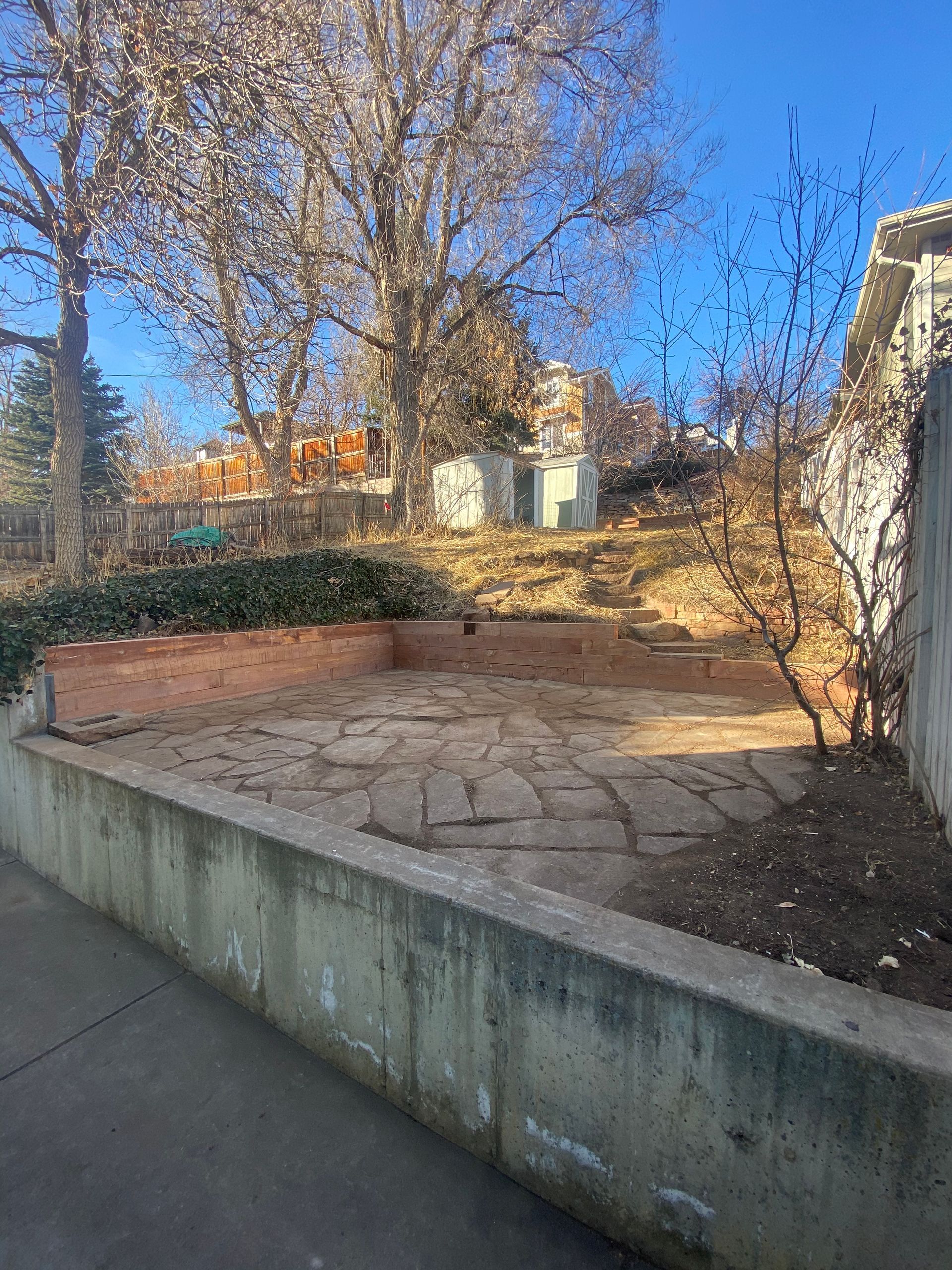 A brick patio with a low wall, situated on a slope with bare trees and a blue sky in the background.