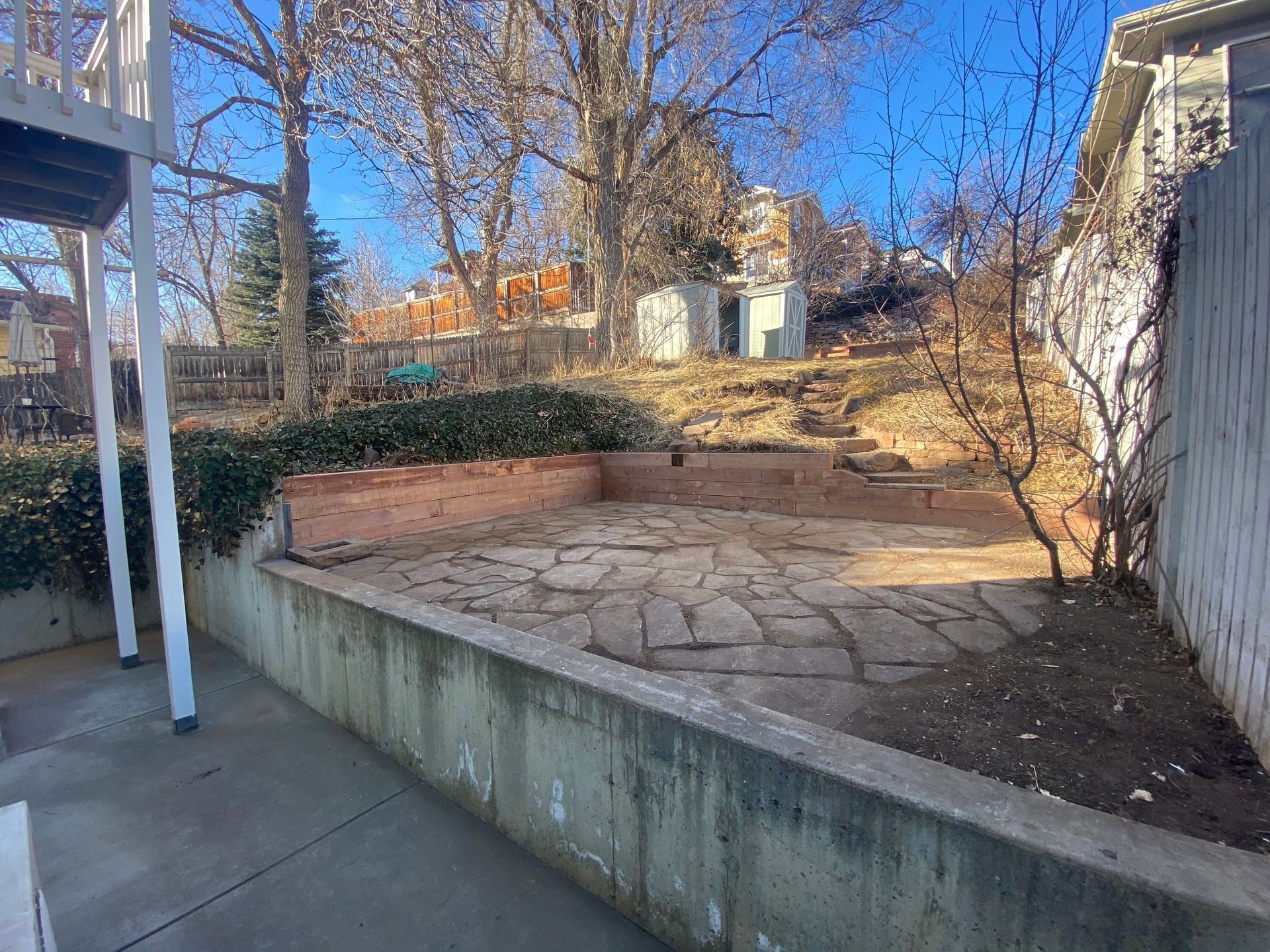 Backyard with a stone patio, retaining wall, and shed on a hillside under a sunny blue sky.