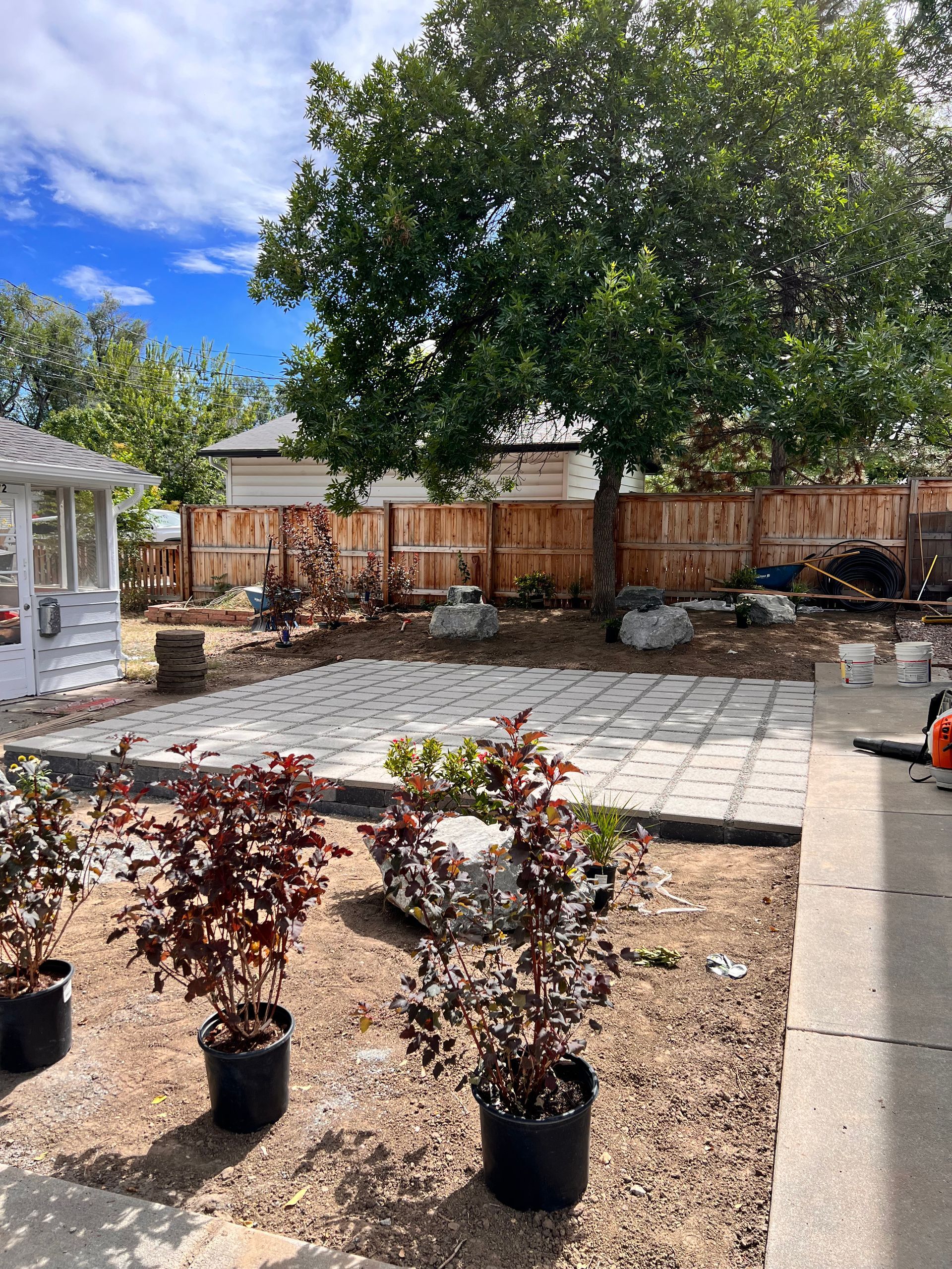Backyard patio under construction with trees, pavers, and potted plants.
