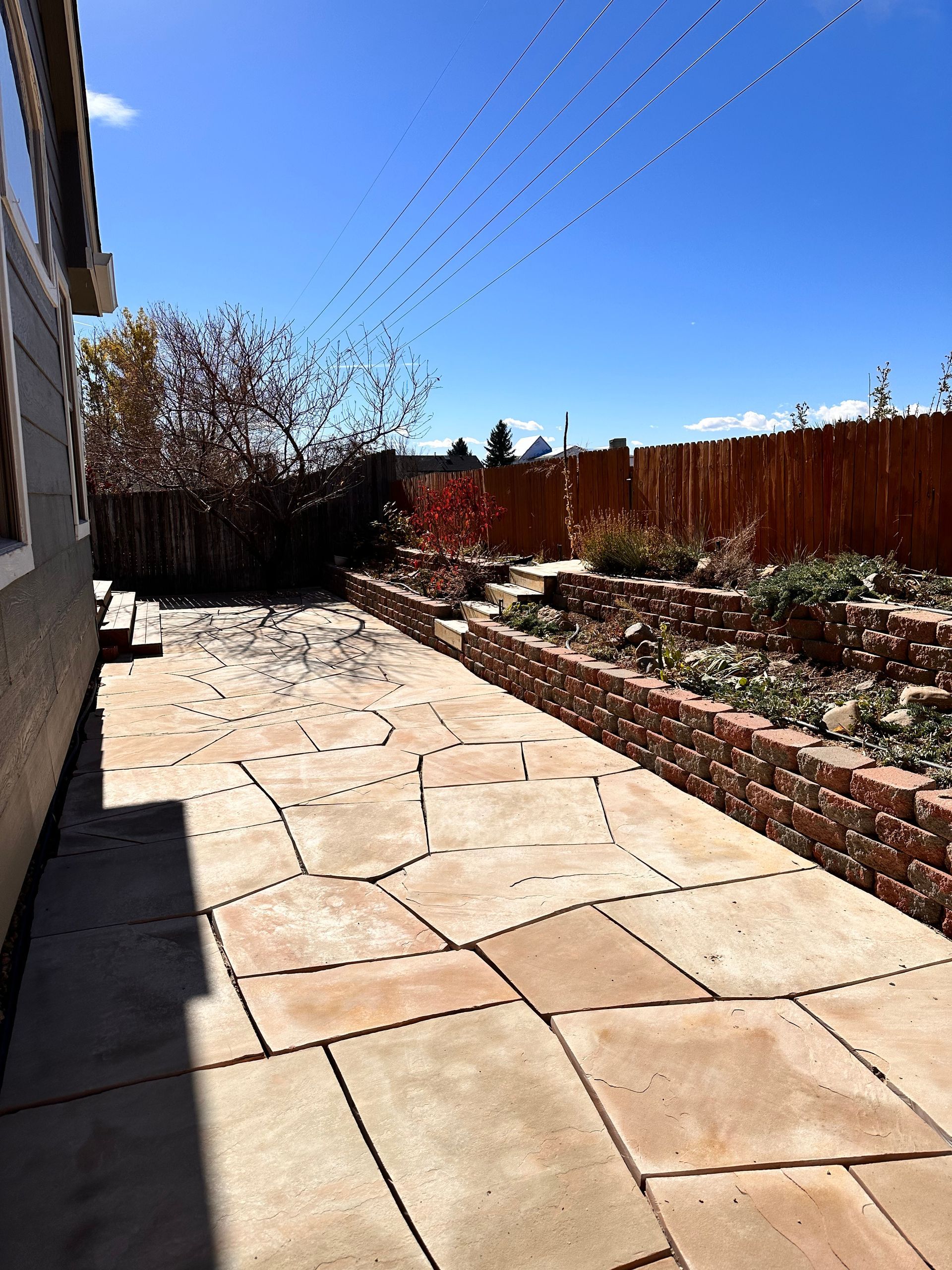 A stone patio next to a building with a red brick planter and bamboo fence under a blue sky.