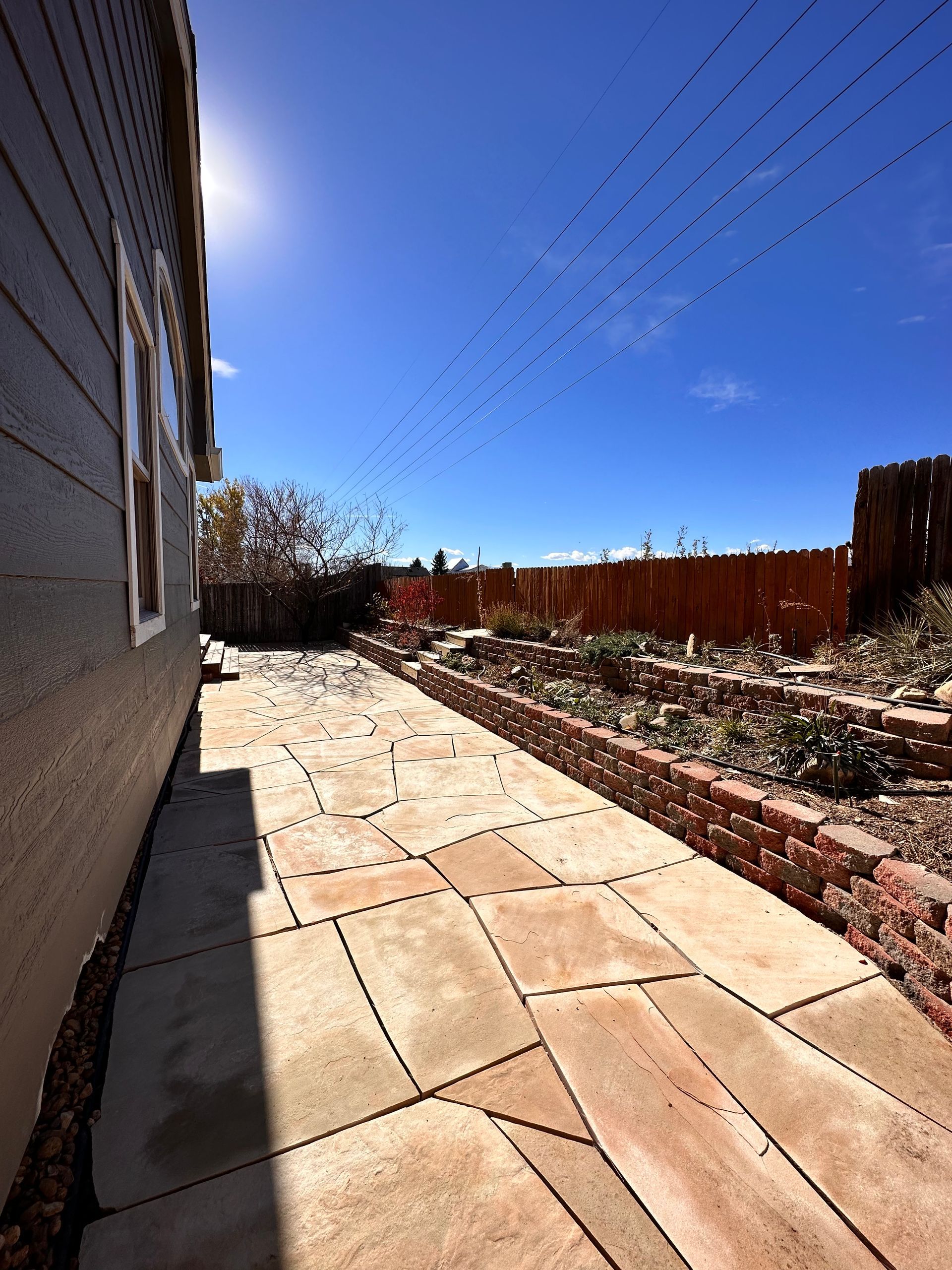 Patio with stone pavers and brick border alongside a dark house, under a blue sky.