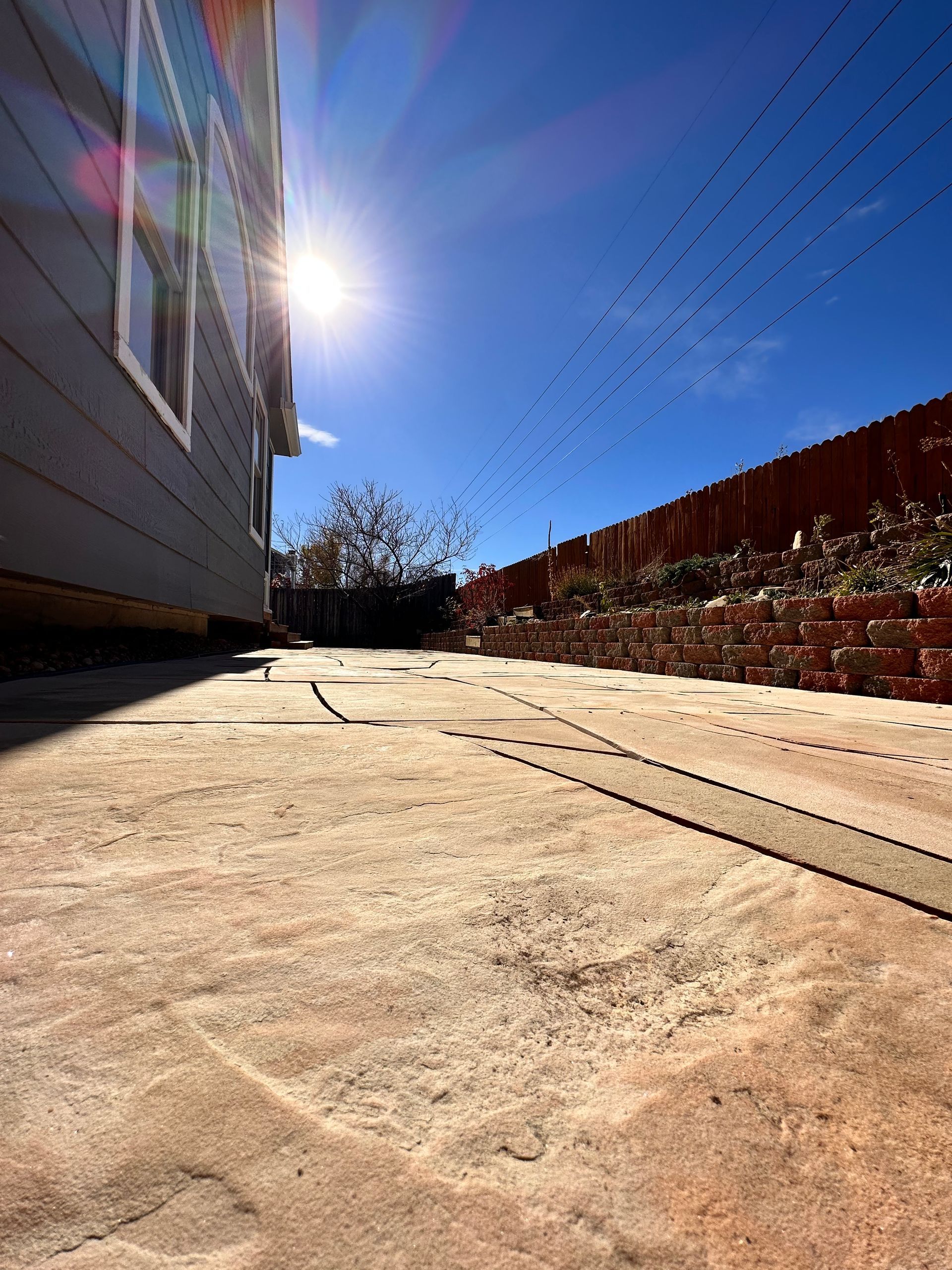 Bright sun shines over a concrete patio next to a gray building and a red brick wall.