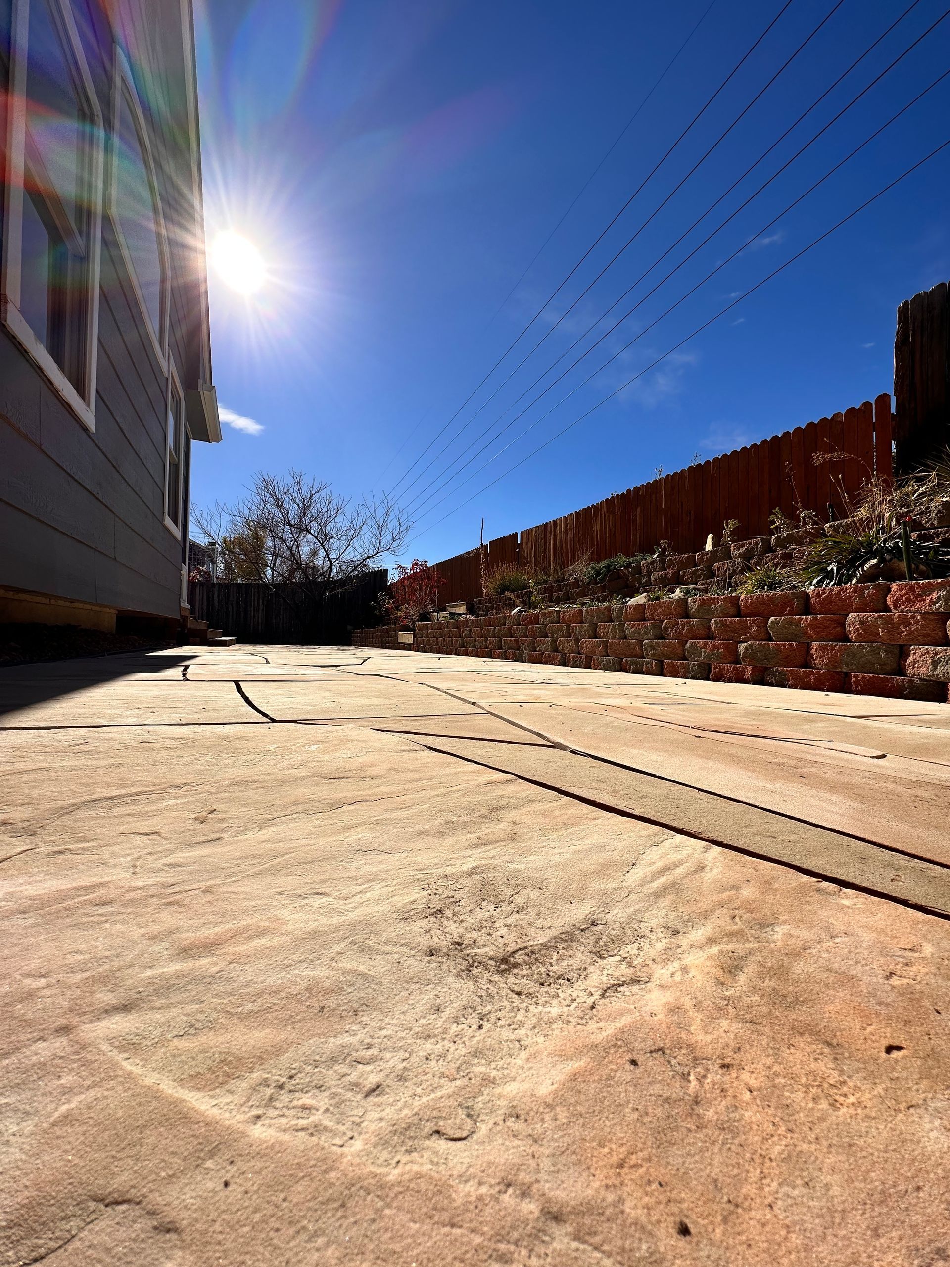 Bright sun over a concrete patio; birds fly against a blue sky, beside a house and fence.