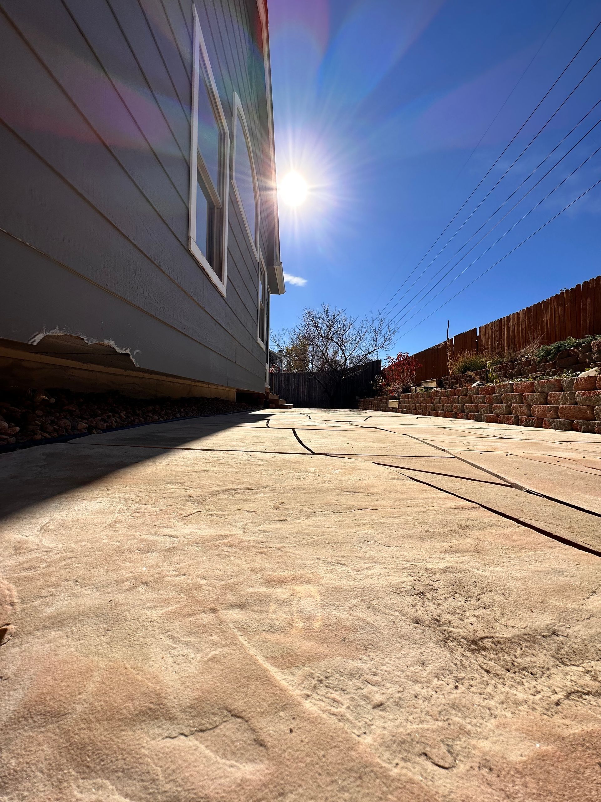 Sunlit, low-angle view of a brick patio beside a gray house. Bright sky, shadow cast across the patio.