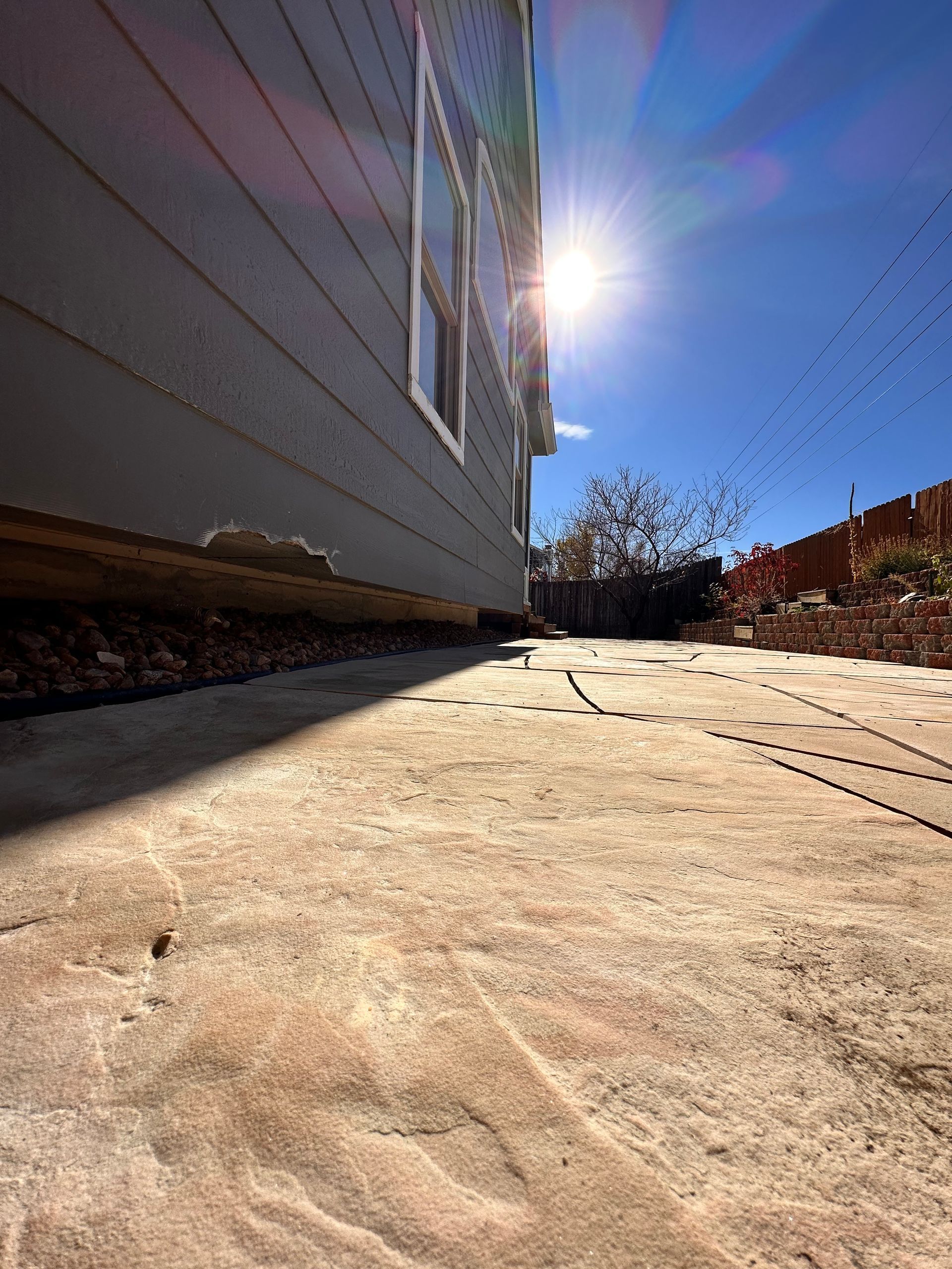 Bright sun over a gray house with a concrete walkway. Birds fly overhead against a clear blue sky.