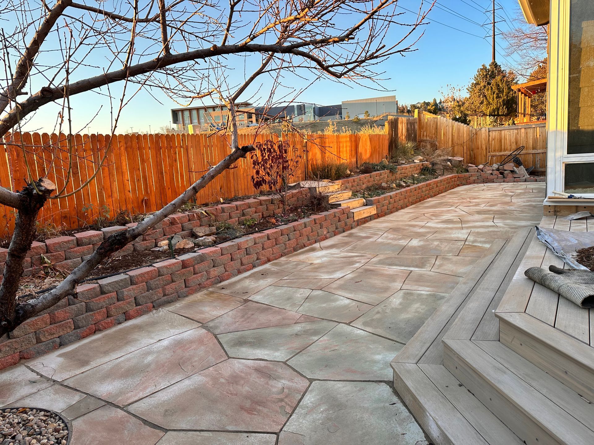 Patio with brick-lined flower beds, wooden fence, and steps leading to a deck in a backyard setting.