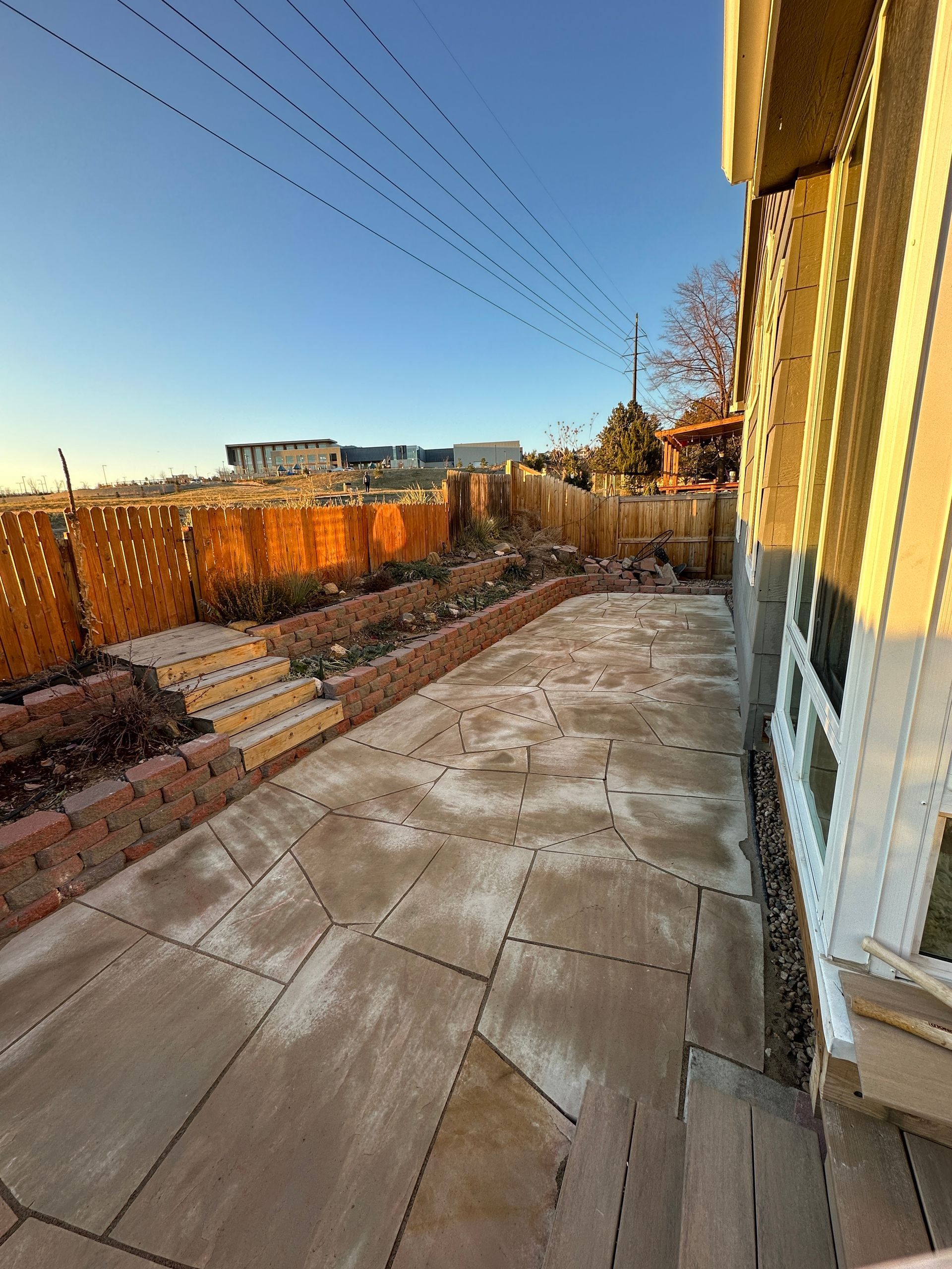 Patio with stone pavers, brick borders, wooden fence, and steps leading to a garden.