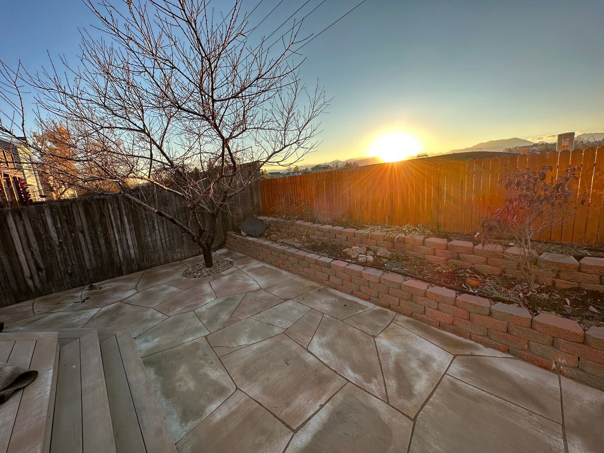 Backyard at sunset with a tree, paved patio, brick wall, and wooden fence.