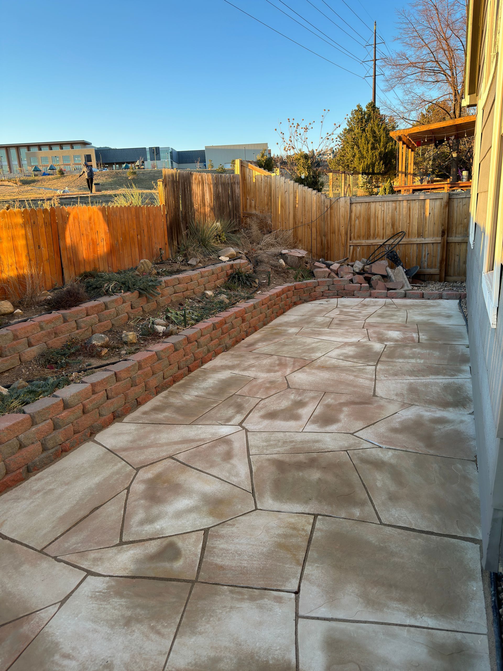 Patio with irregular stone paving, brick retaining wall, wooden fence, and sparse landscaping.
