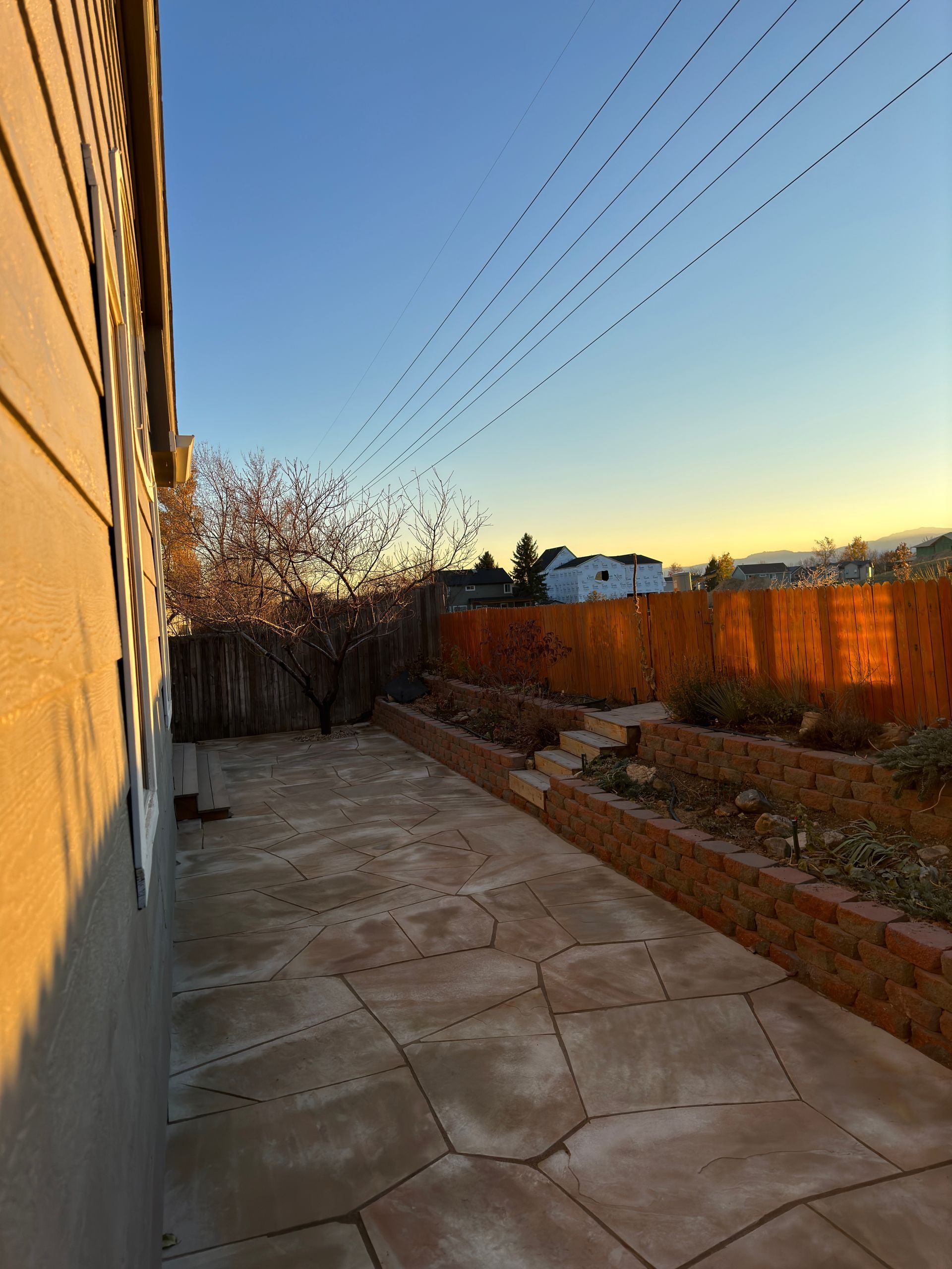 Stone patio next to a beige house, leading to a garden with a sunset sky.