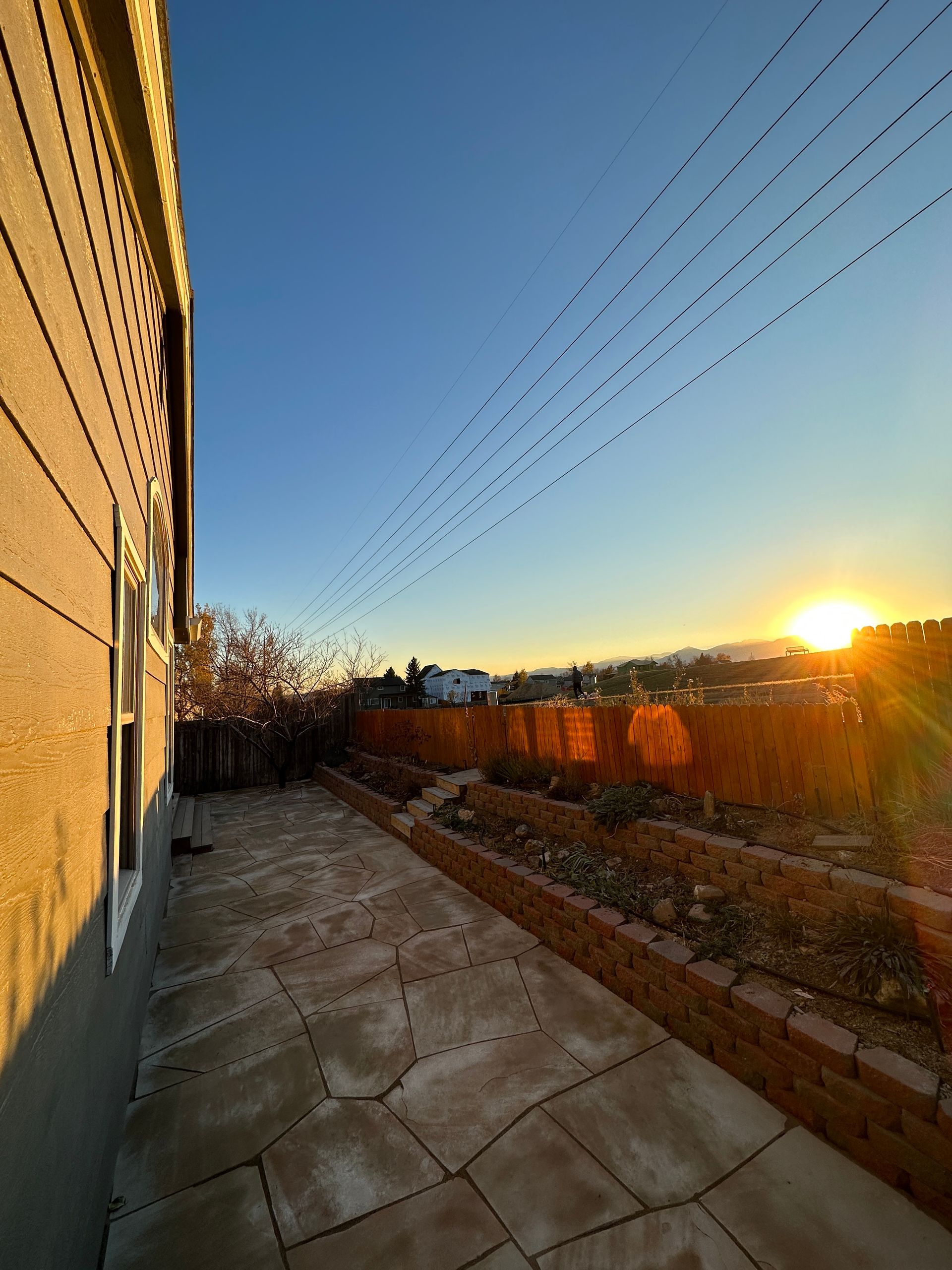 Patio with brick-lined garden beds at sunset, side of house on the left, birds in the sky.