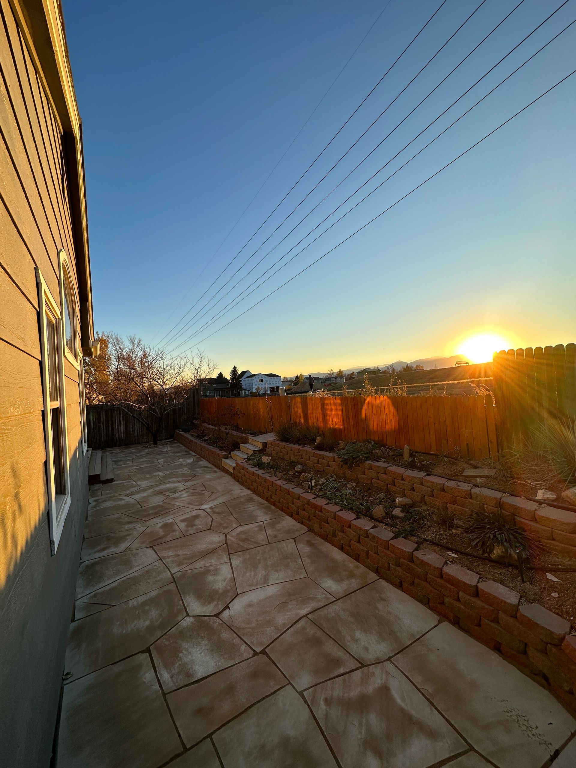 Patio with stone pavers, sun setting over a wooden fence and distant hills under a blue sky.