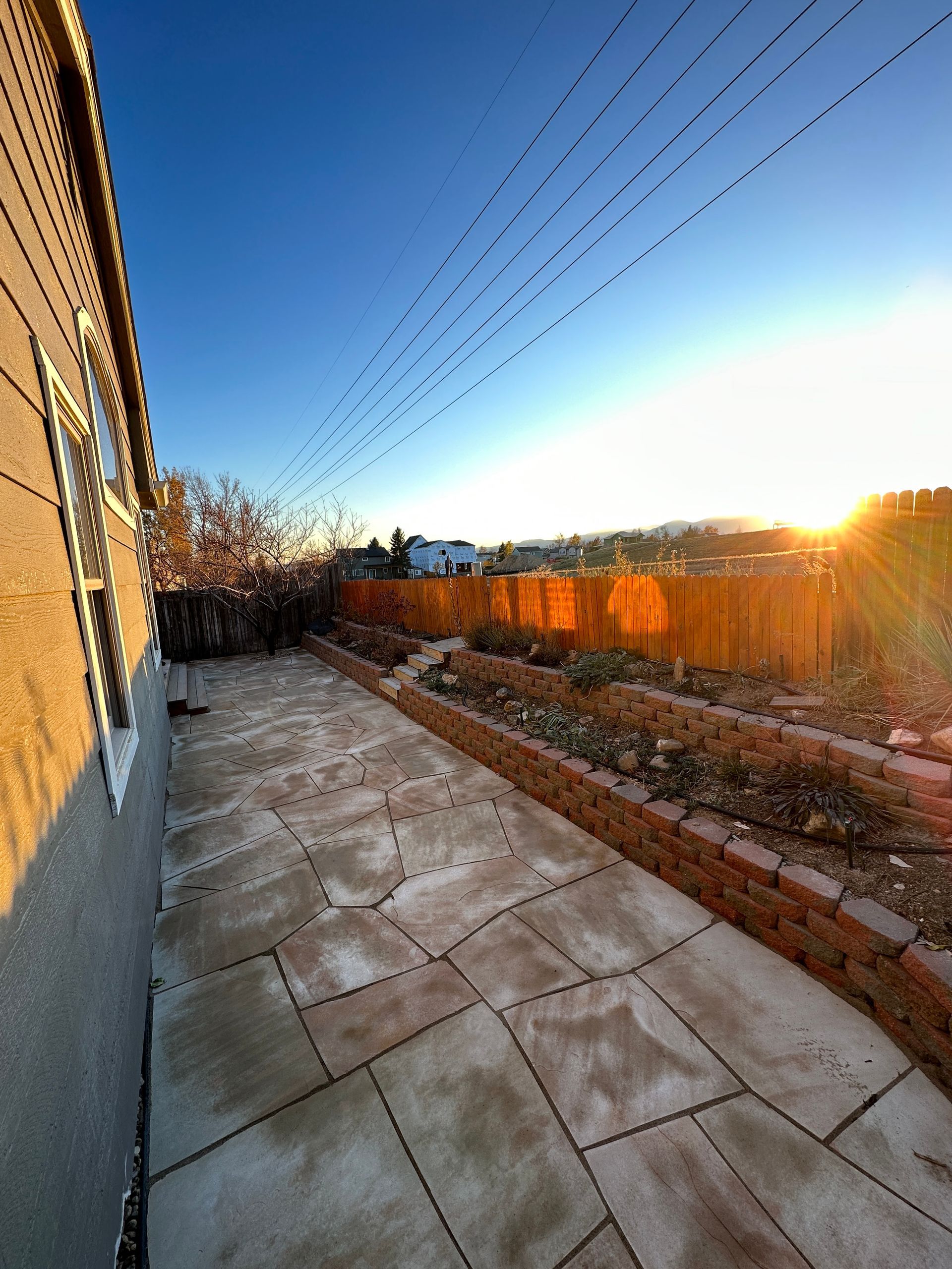 Stone patio and raised beds along a wall, with sun setting over a wooden fence and distant hills.