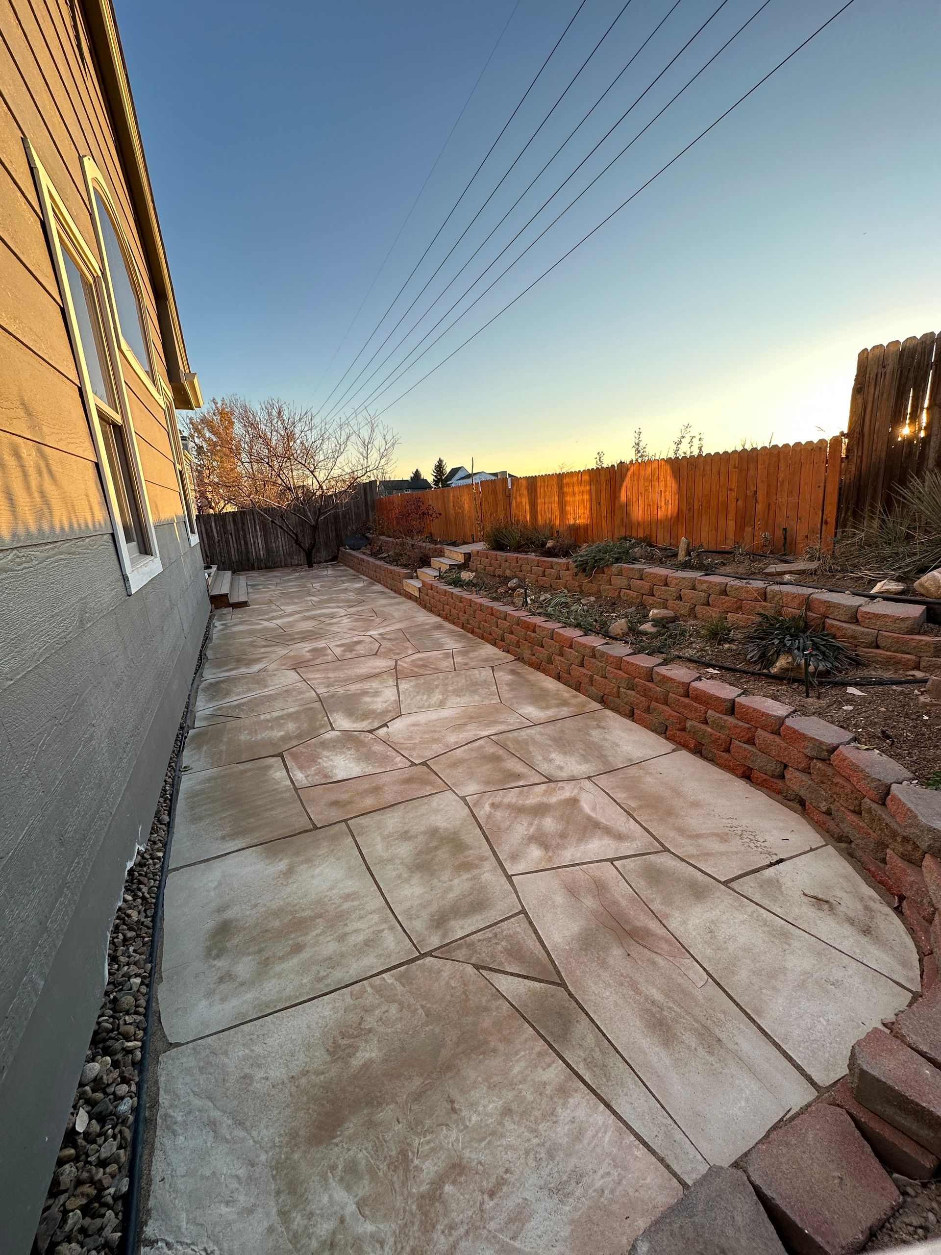 Stone patio beside a house with a brick border, overlooking a garden and fence at sunset.