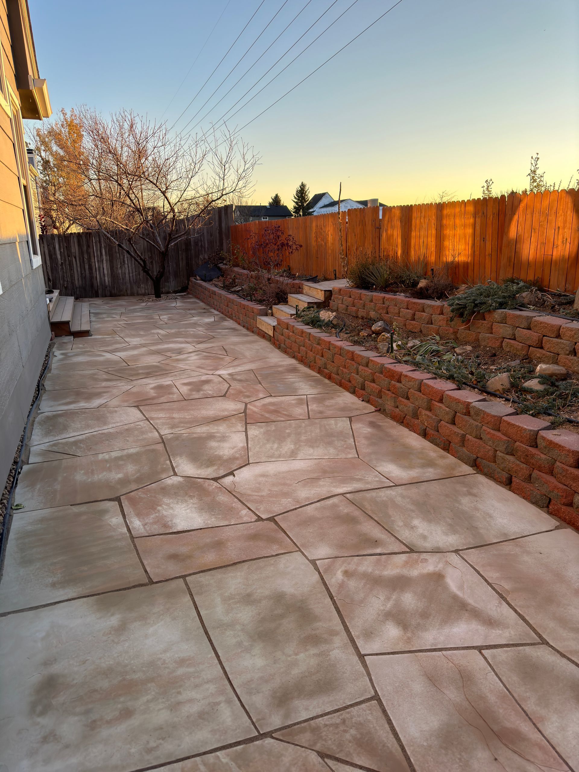 A backyard patio with stone pavers, brick planters, and a wooden fence at sunset.