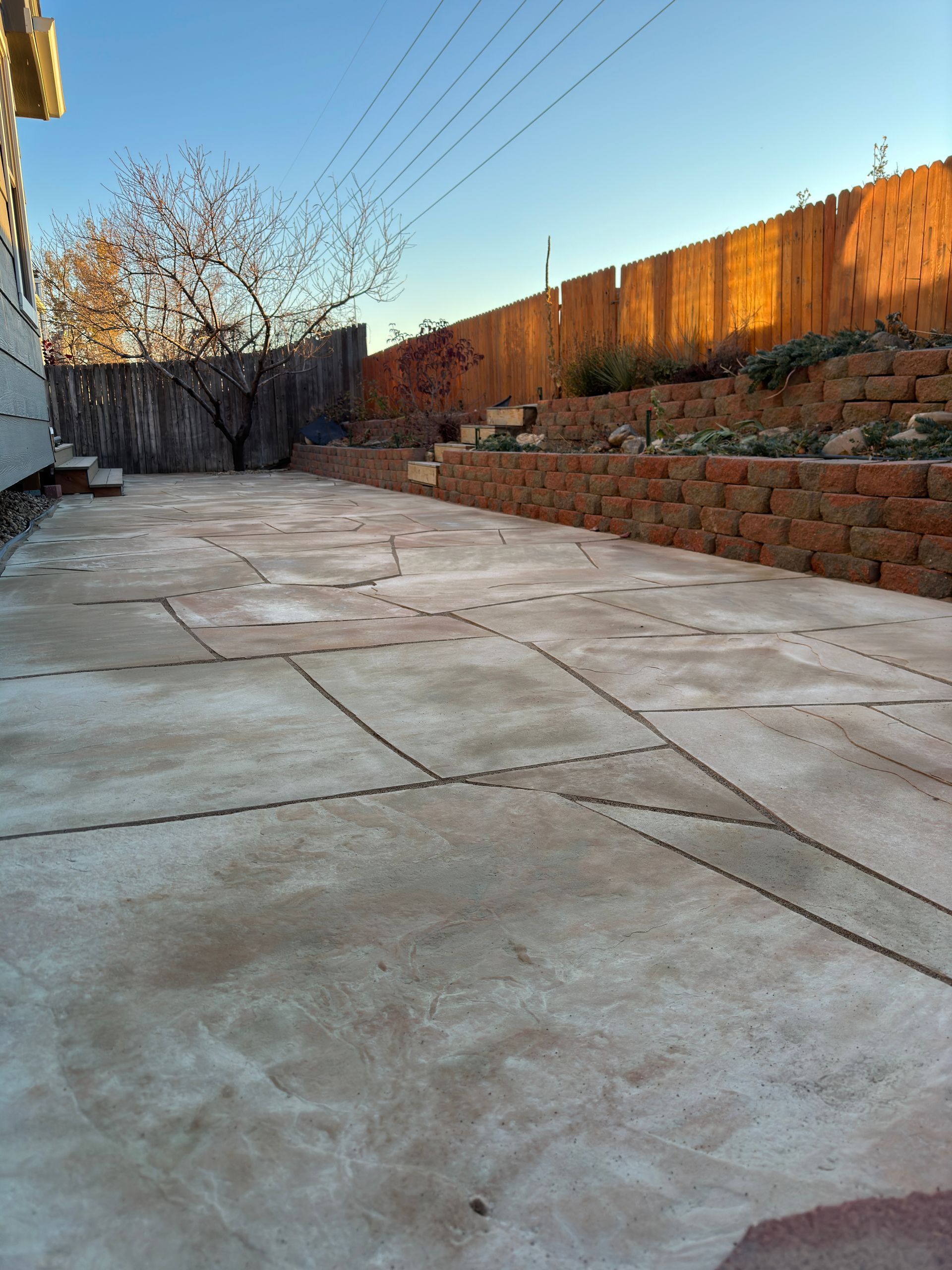 Large concrete patio next to a brick retaining wall and wooden fence, with a tree and blue sky in the background.
