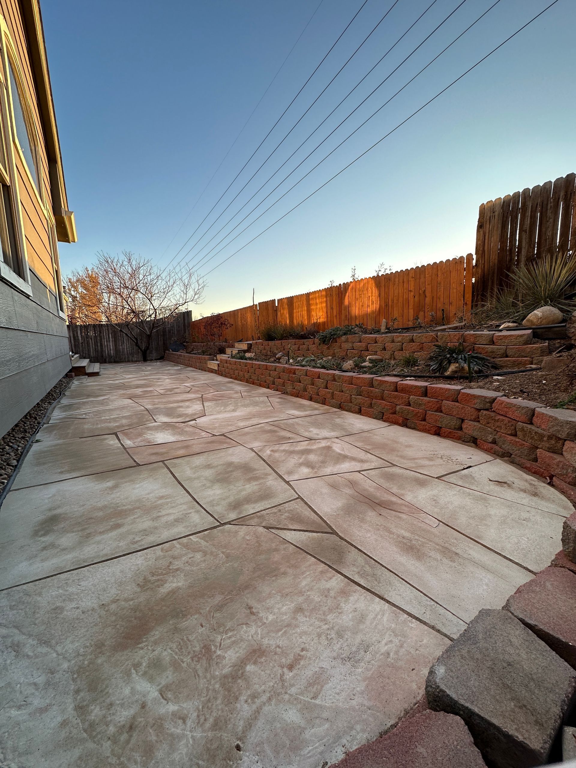 Concrete patio with brick retaining wall in a backyard with a wooden fence and clear sky.