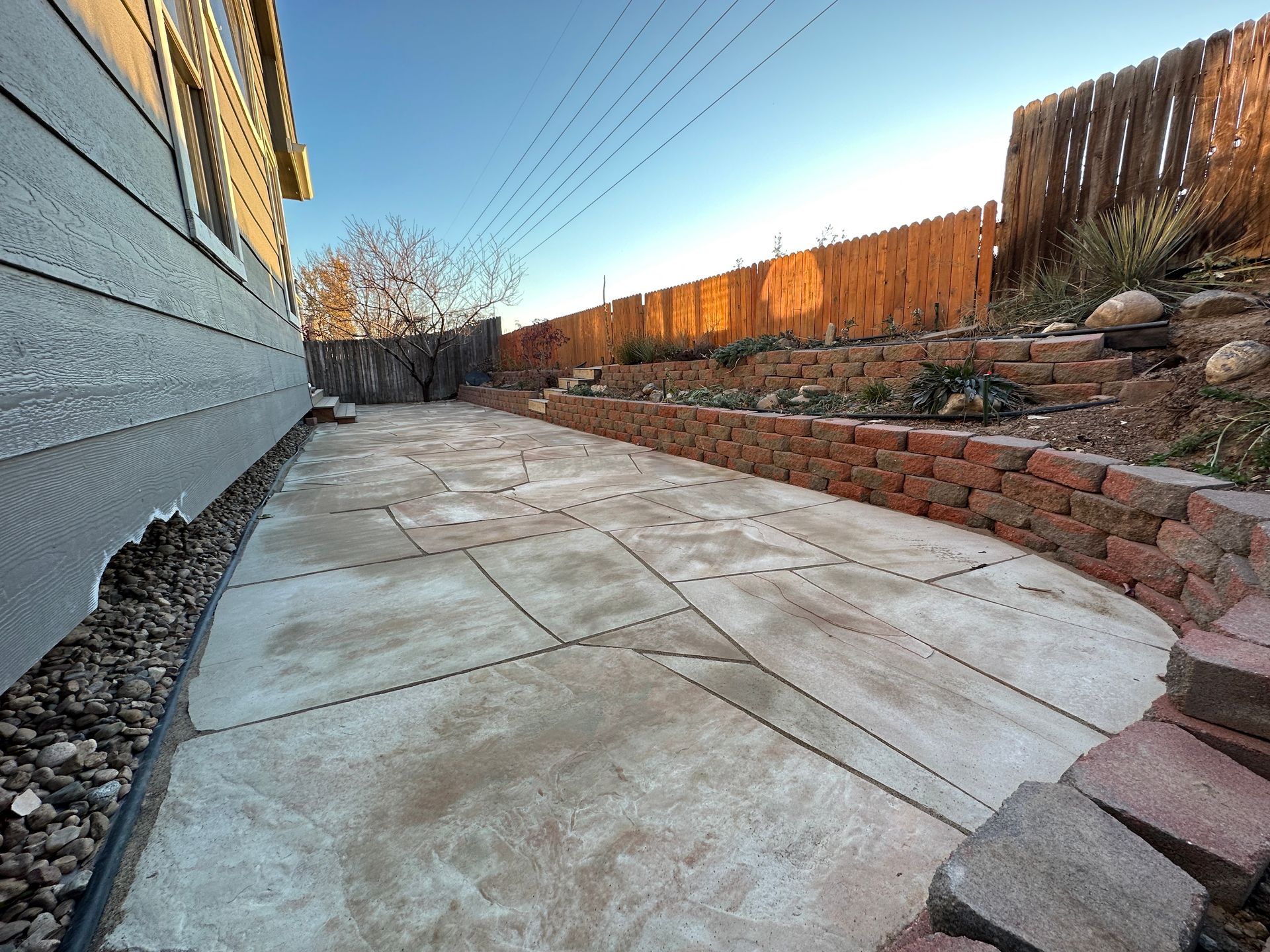 Concrete patio alongside a gray house, bordered by a brick wall and wooden fence, under a blue sky.