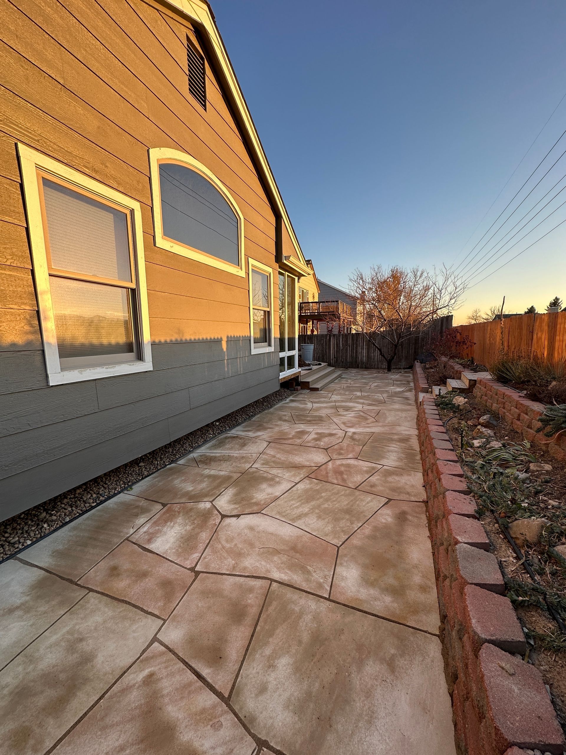 Side view of a house with a concrete patio and a garden bed, lit by the setting sun.