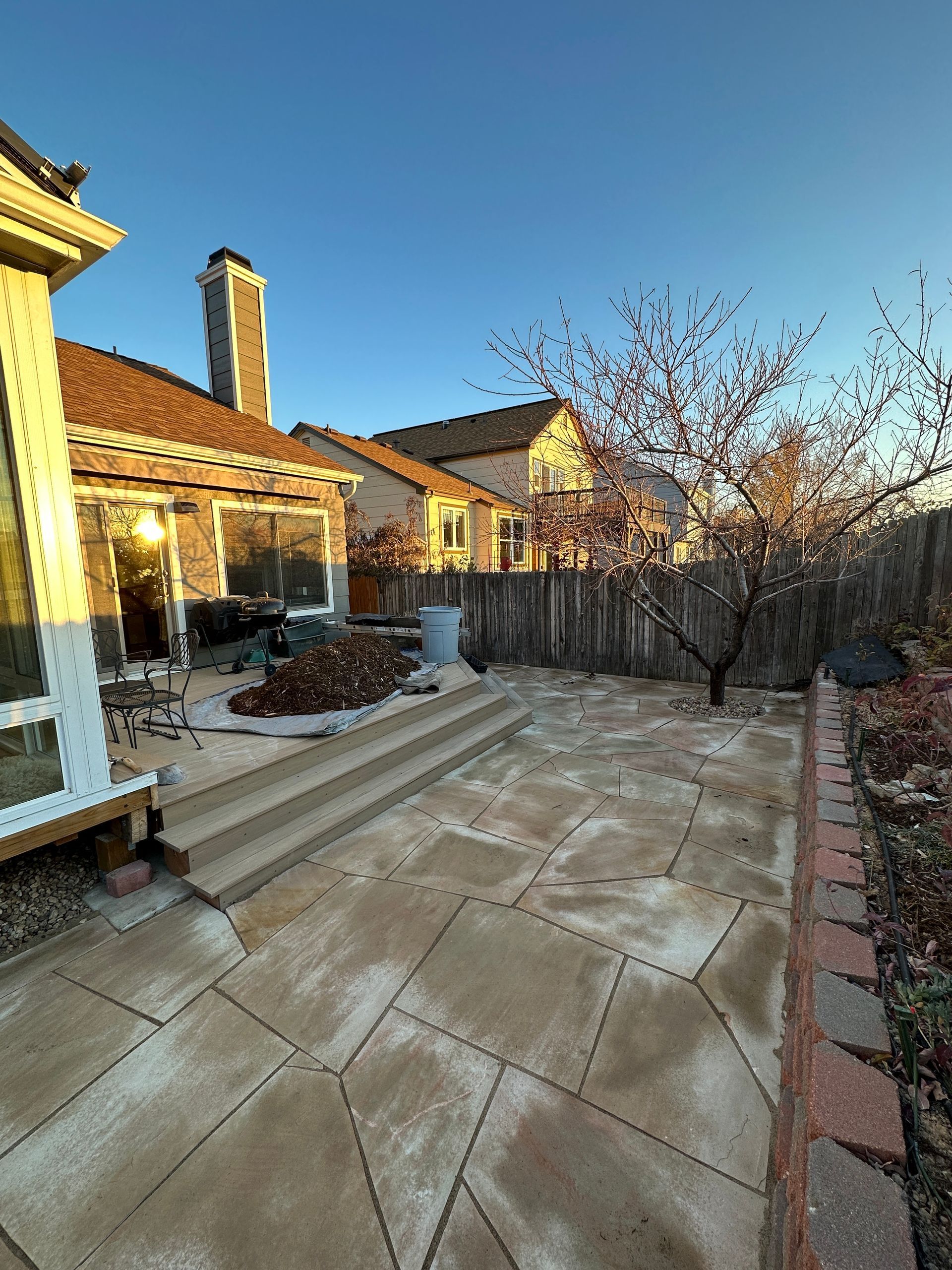 Backyard patio with stone paving, steps, and a tree, under a clear blue sky.