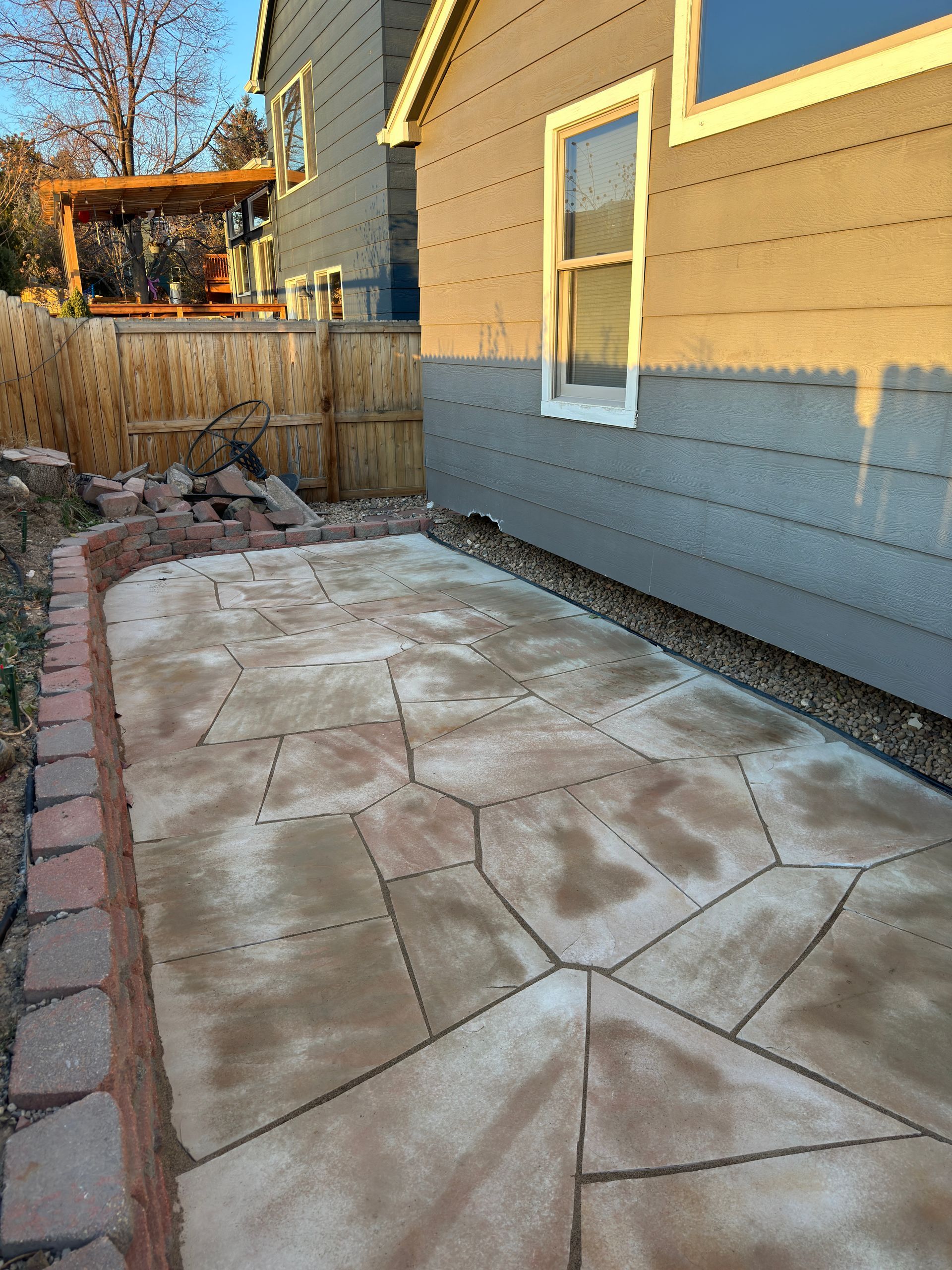 Flagstone patio with brick edging beside a gray house with a wooden fence.