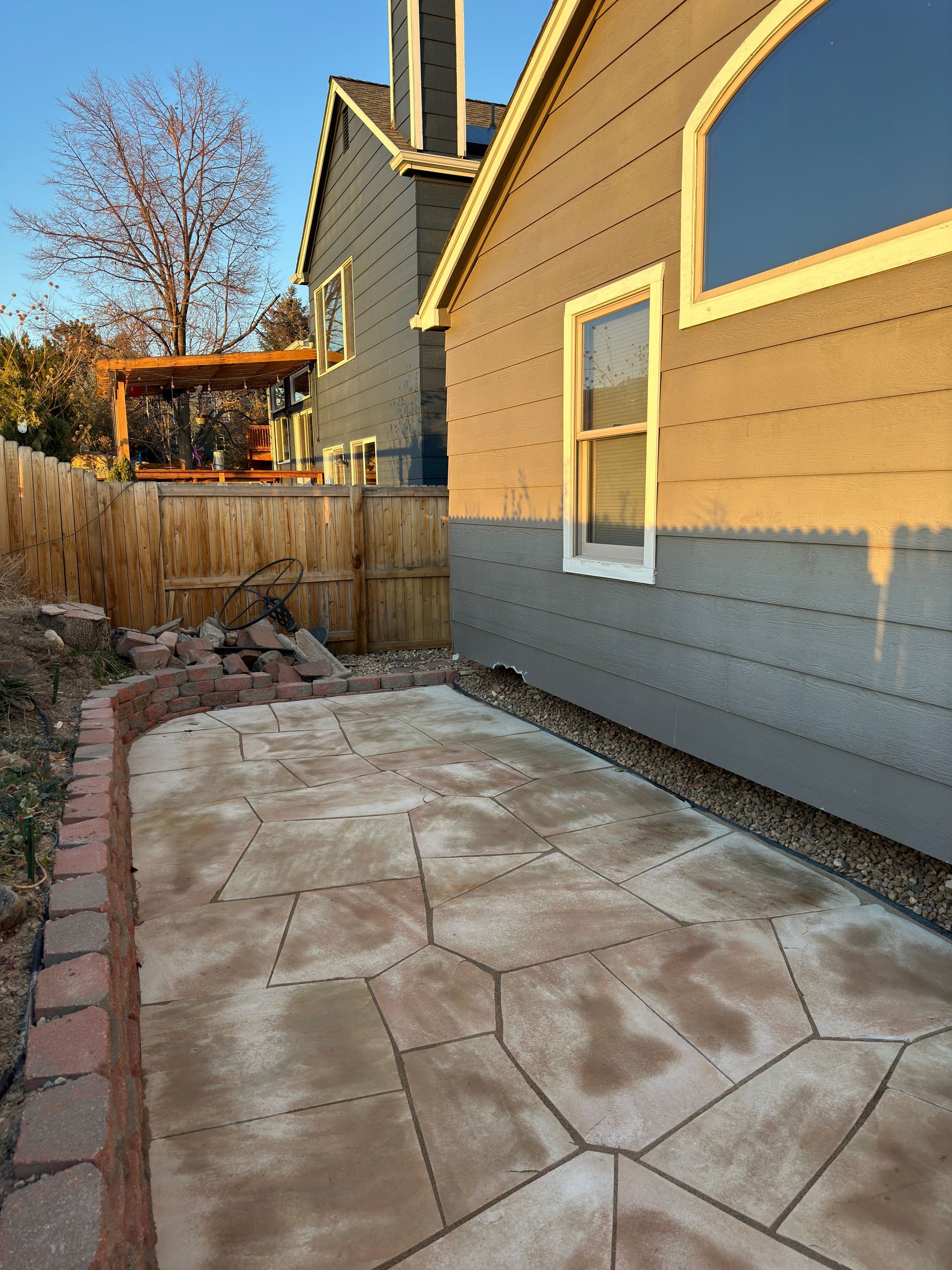 Stone patio next to a gray house, bordered by red bricks and a wooden fence, with a pergola in the background.