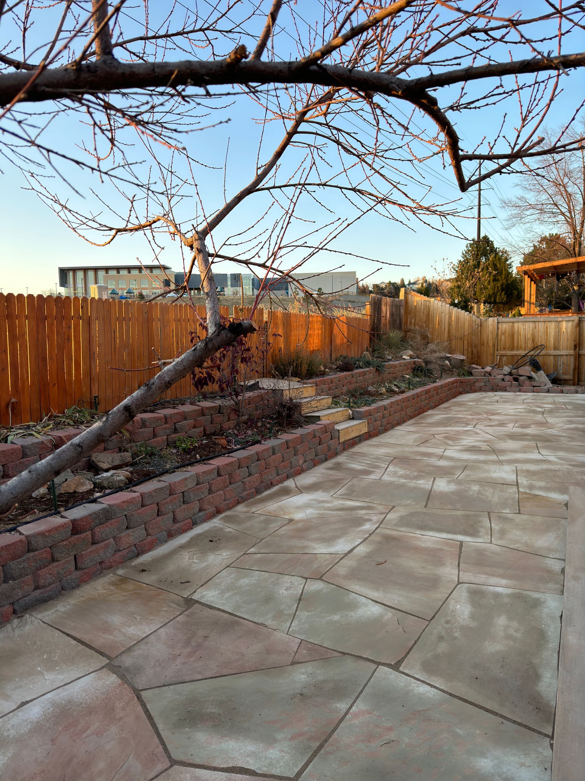 Backyard with concrete patio, brick retaining walls, wooden fence, and bare tree branches against a blue sky.
