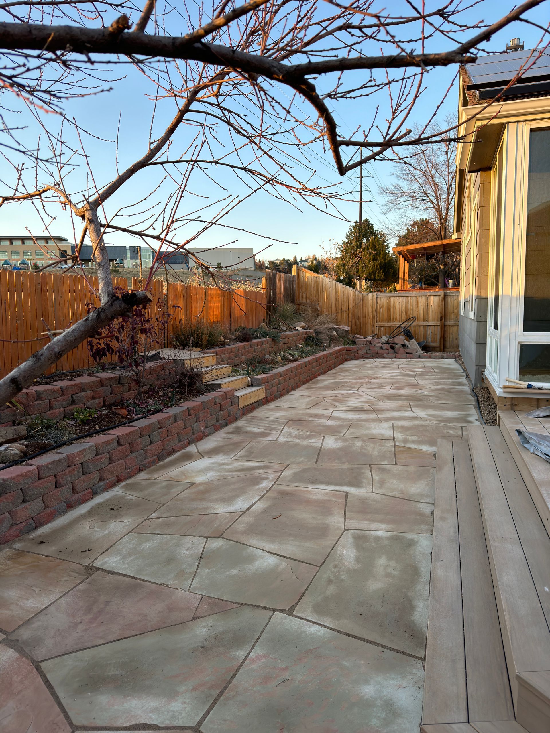 Patio with stone pavers, red brick retaining walls, and wooden fence. Bare tree branches in the foreground.