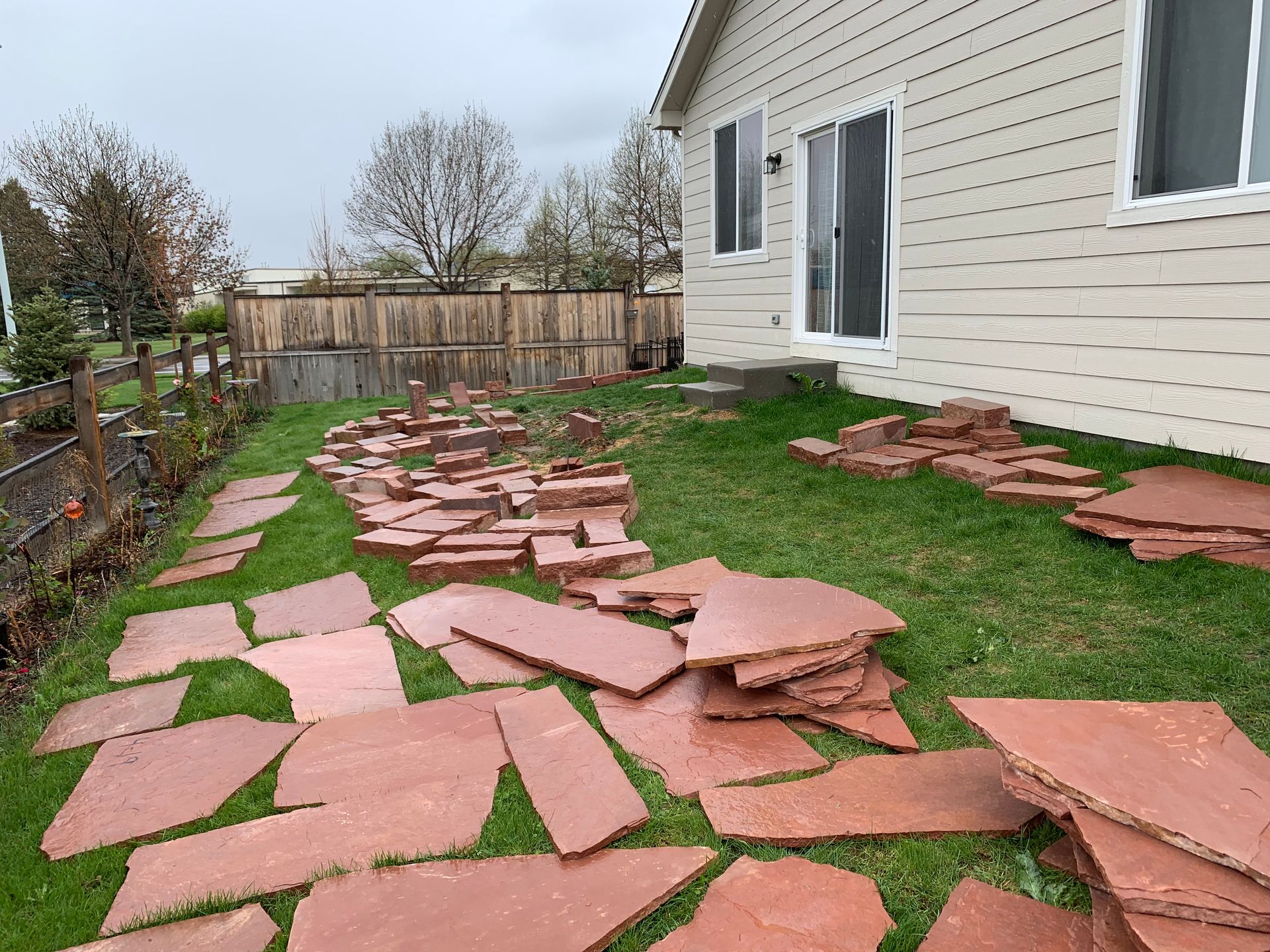 Red stone path construction in a backyard with a beige house, green grass, and wooden fence.